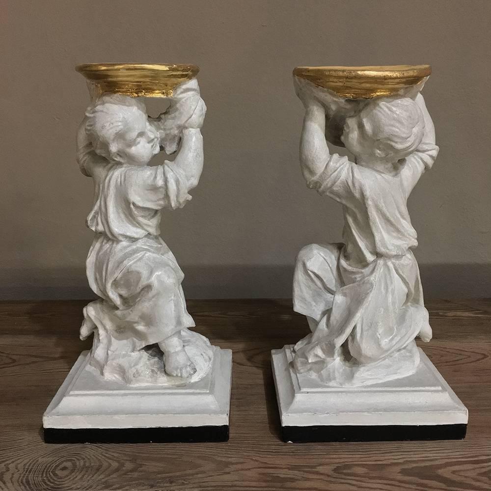 Pair of 19th Century Italian Painted Statues of Cherubs feature a mirrored image effect, with the cherubs depicted in a kneeling stance holding up a golden bowl. The painted finish on carved wood has achieved a lovely distressed patina over the