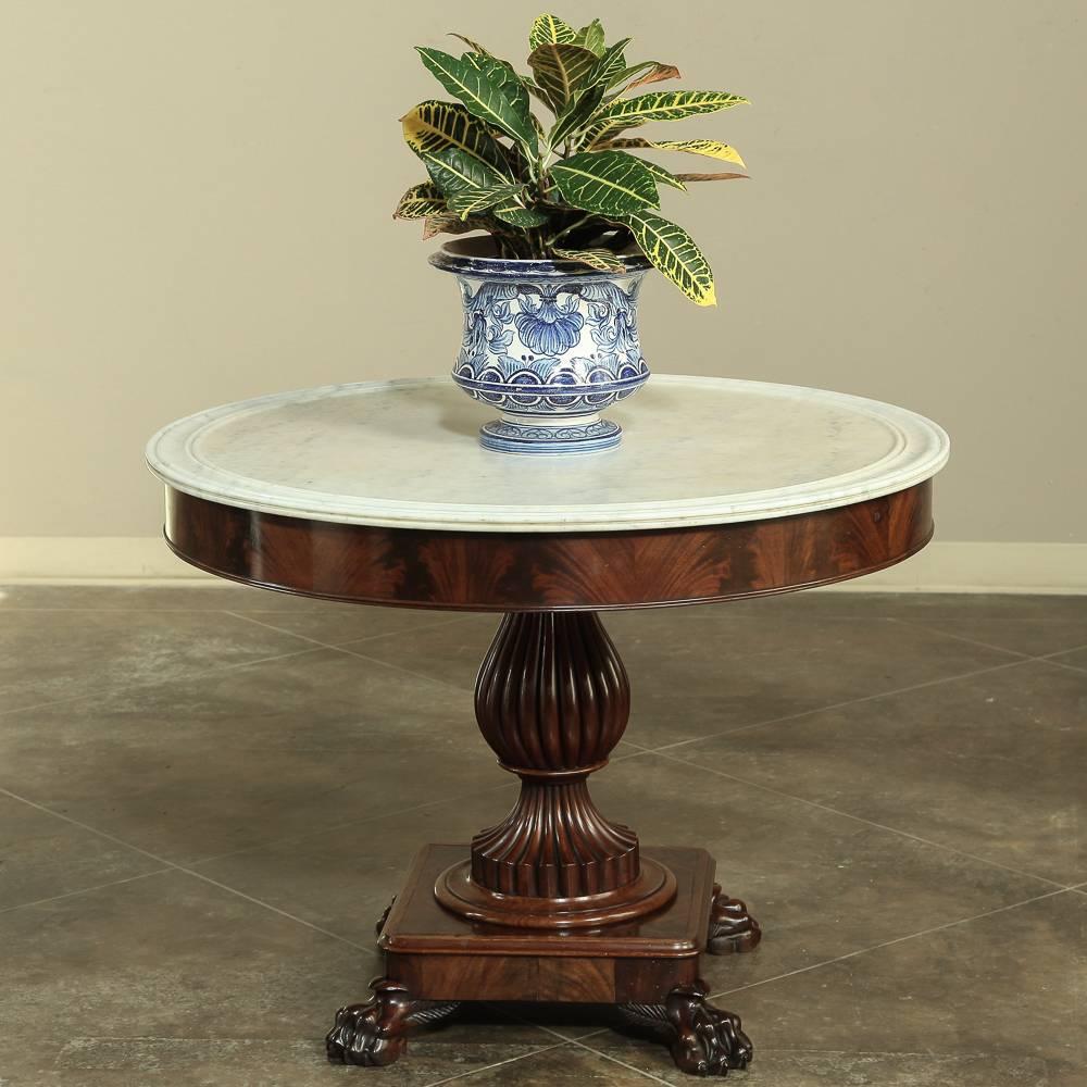 19th century Louis Philippe marble-top center table was handcrafted from exotic imported mahogany and features bold lions paw feet, crotch mahogany veneered apron, a fluted urn pedestal, and the original finger groove Carrara marble top! Designed to