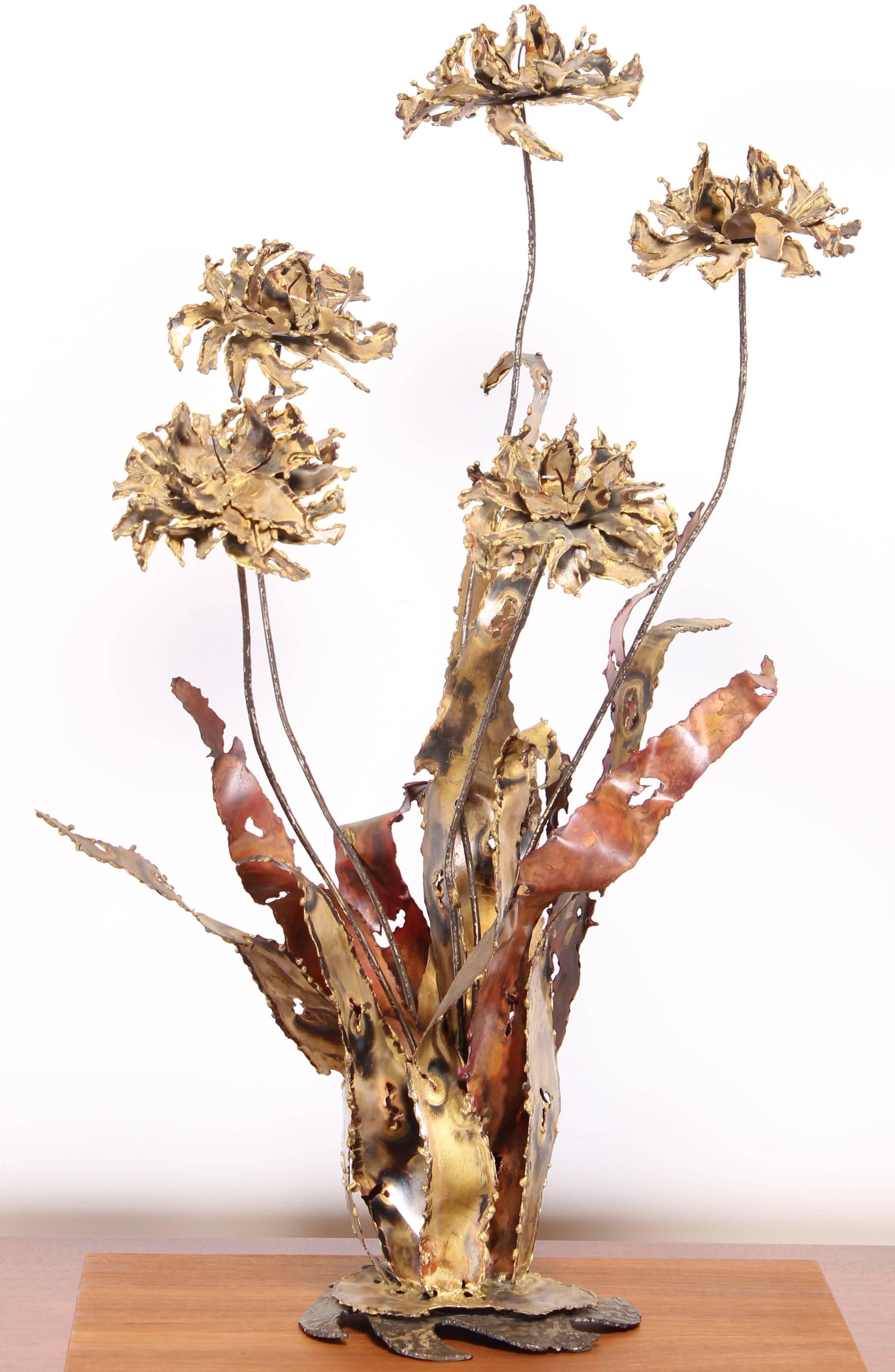 A sculpture with torch cut strips of brass metal that form the leaves and flower petals creating a Brutalist abstract flower. Bottom is signed Silas Seandel.