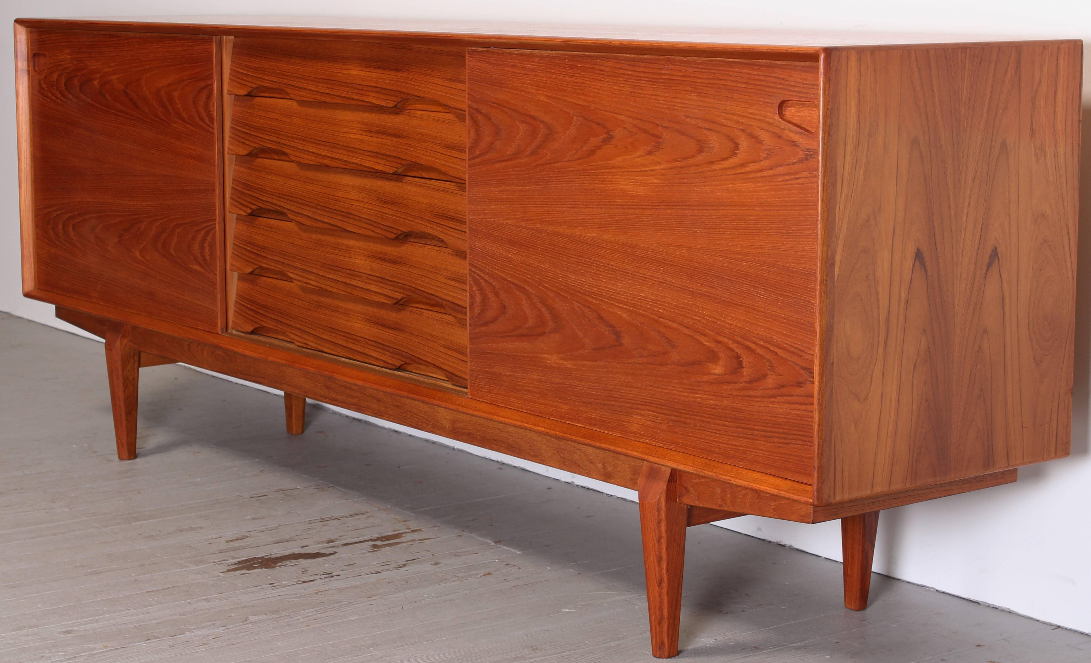 Teak credenza made by Dyrlund of Denmark. Two sliding doors and five drawers, top drawer has green felt lining. Adjustable shelves, use none or adjust the three included shelves as needed. Dyrlund label affixed inside cabinet. Pair are available. 
