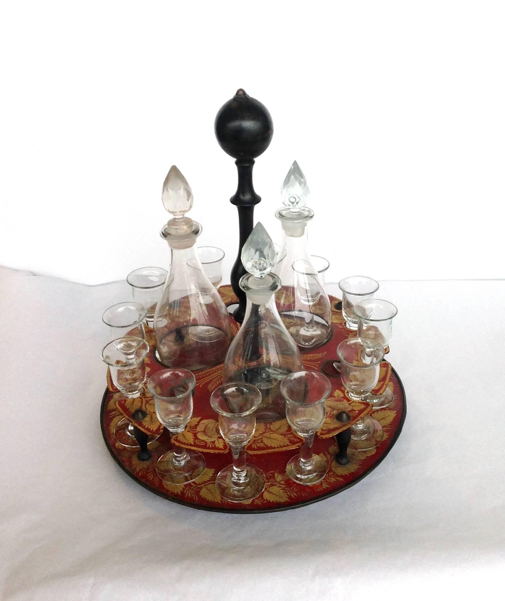 Regency red and gold tole decanter set, red with gilt decorations on swivel base. It has a set of three decanters and 12 liqueur glasses. Two glasses have been replaced and one decanter has a crack.