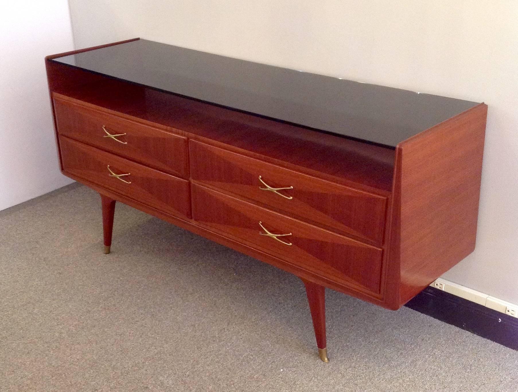 Mid-Century Italian fruitwood console table with a floating black glass top and subsidiary shelf over two-tone mahogany, four-drawer base. It has a modern sleek design with brass handles and tapered legs highlighted by brass sabots.