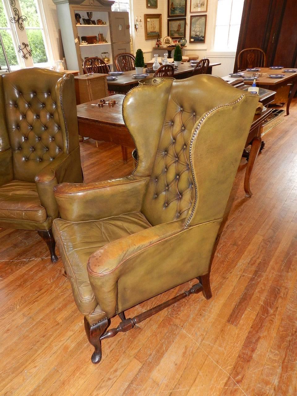 A pair of antique buttoned leather wing chair design club chairs with oak cabriole legs and stretchers. Both chairs have out swept arm rests and shaped seats. The buttoned leather upholstery exhibits the beautiful aged patina that can only be found