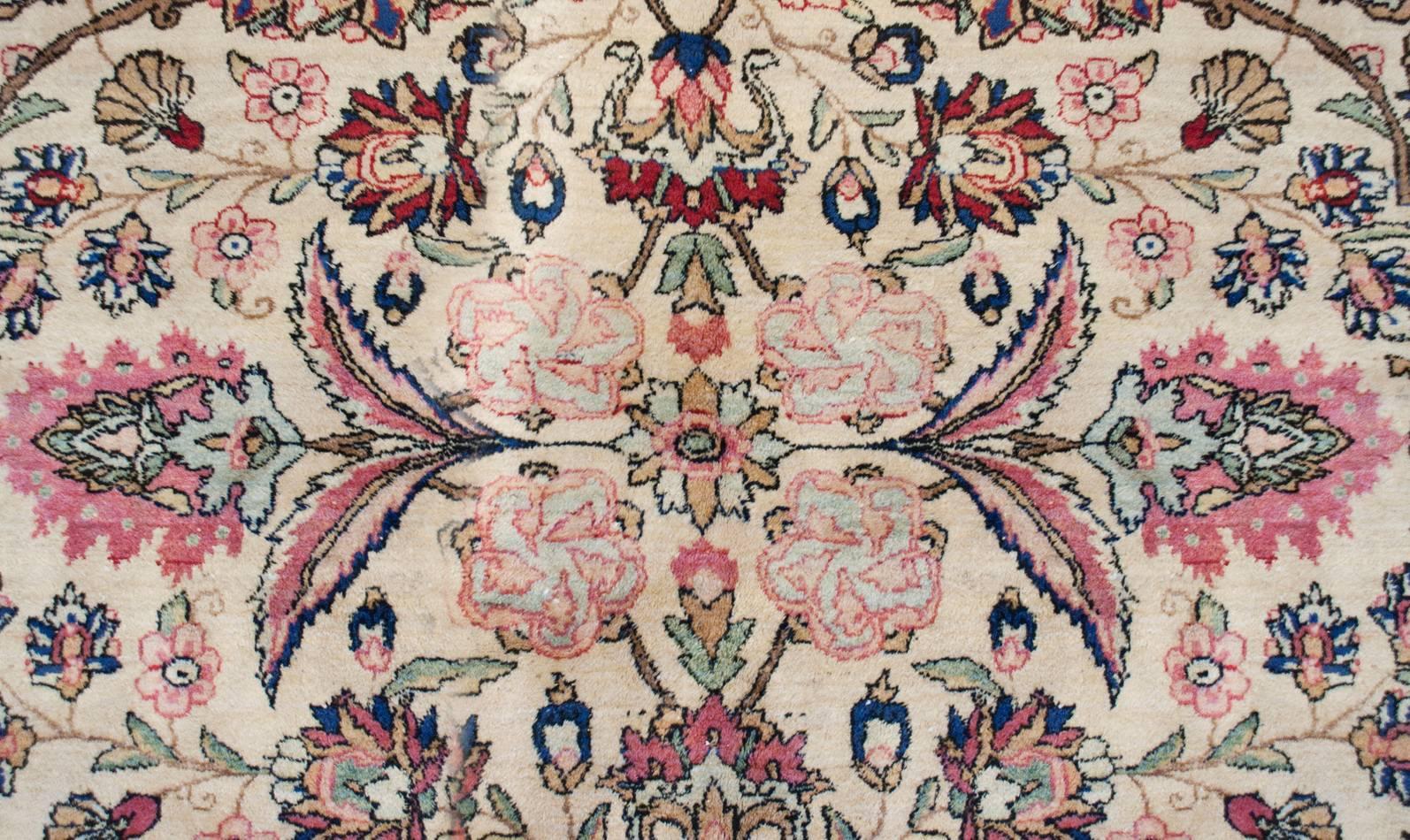 An early 20th century Persian Kirman rug with an intensely woven multi-colored mirrored pattern of scrolling vines and myriad floral varieties on a cream colored background. The border is wide comprised of one wide floral border on a crimson