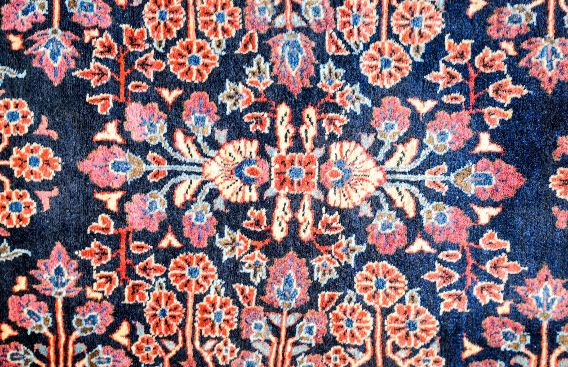 A Classic and wonderful early 20th century Persian Sarouk Mohajeran rug with a bold but traditional mirrored floral pattern woven in crimson, pink, light indigo, and gold, on a dark indigo background. The border is contrasting with a wide cranberry