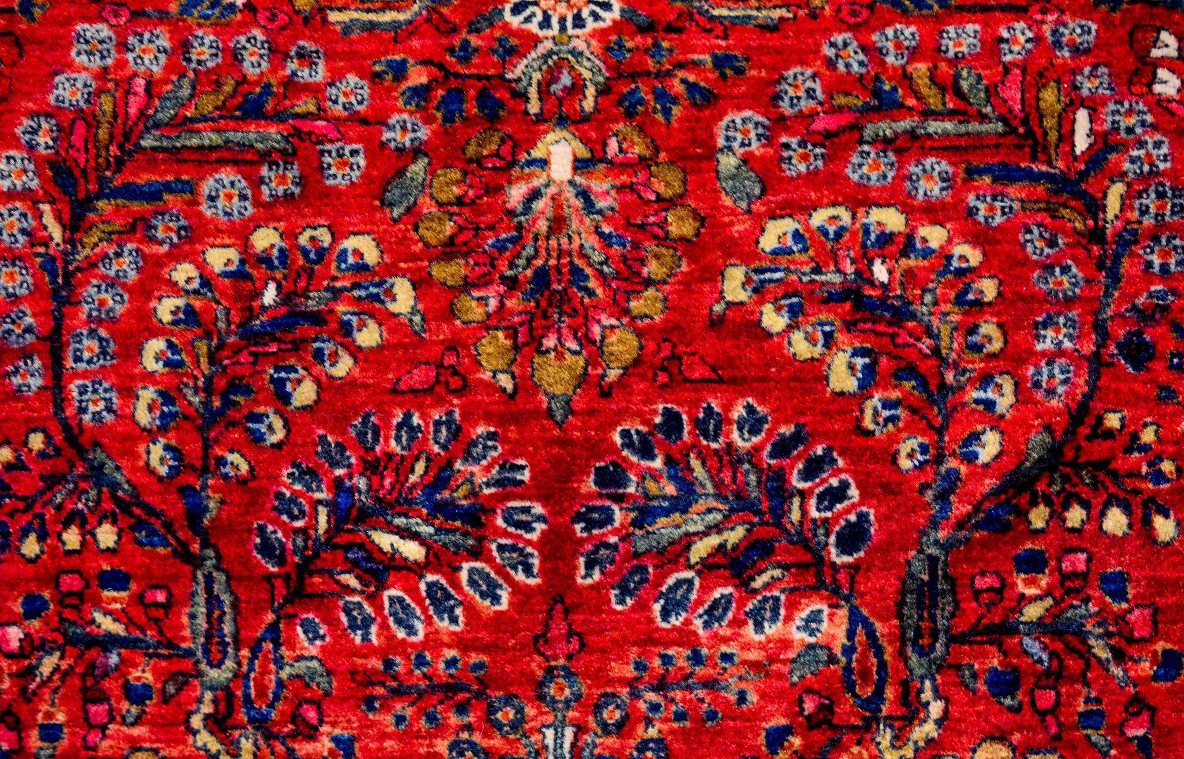 A beautiful early 20th century Persian Sarouk rug with a traditional all-over mirrored floral and vine tree-of-life pattern woven in light and dark indigo and cream colored wool, on a rich cranberry background. The border is complex with a wide