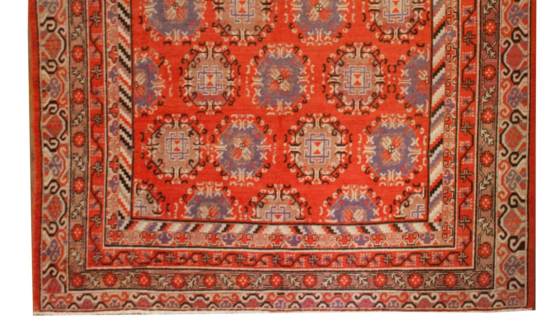 An early 20th century Central Asian Khotan rug with multiple geometric medallions of alternating pale and dark lavender, creating diagonal stripes across the field, all on a rich rusty red background. The border is comprised of multiple thin borders