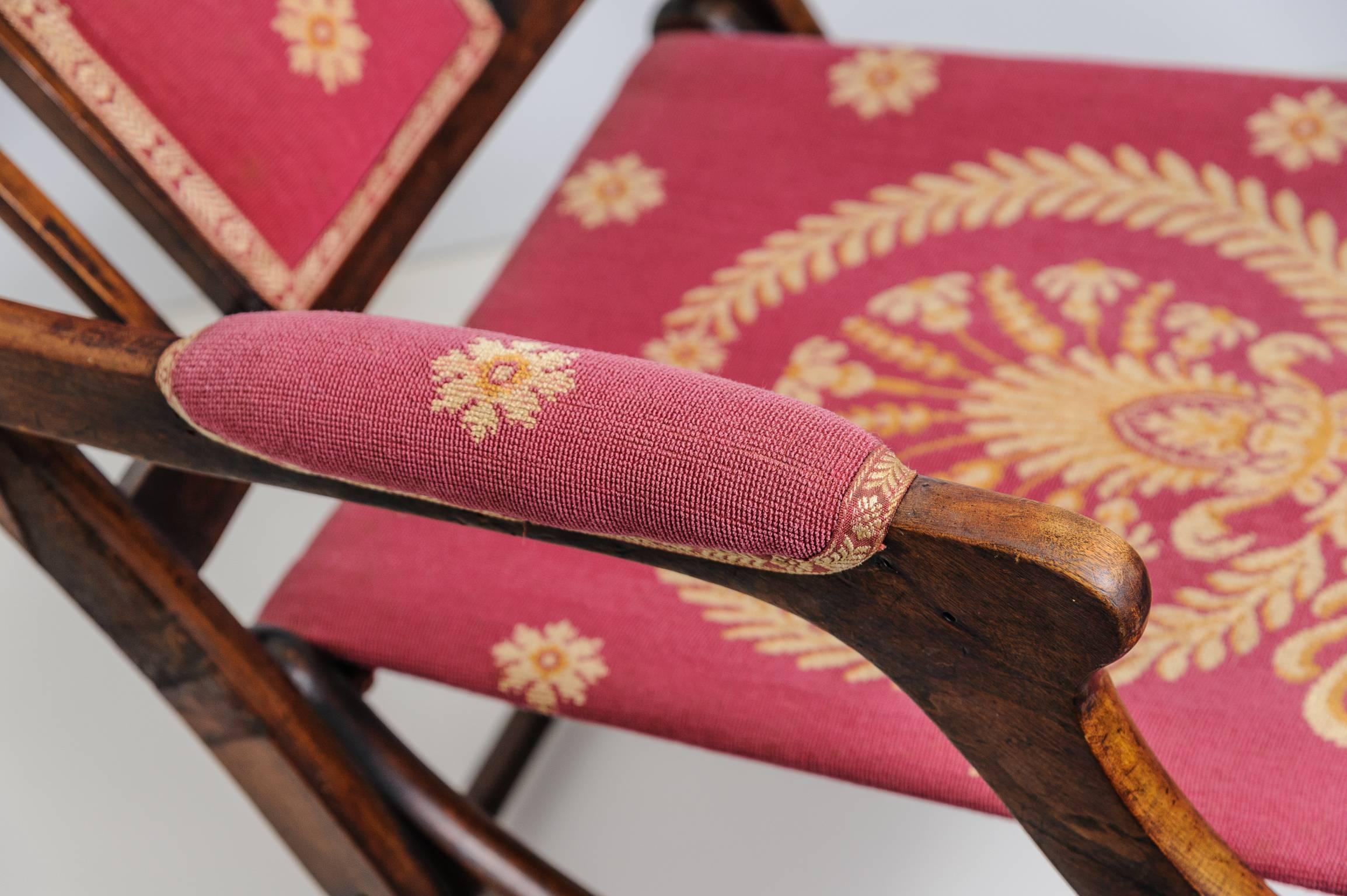 This 19th century campaign style chair was purchased in France and is made of walnut. The woven upholstery fabric is a gros point technique in a muted salmon red and two-tone gold coloration. The central seat medallion is a stylized crown with
