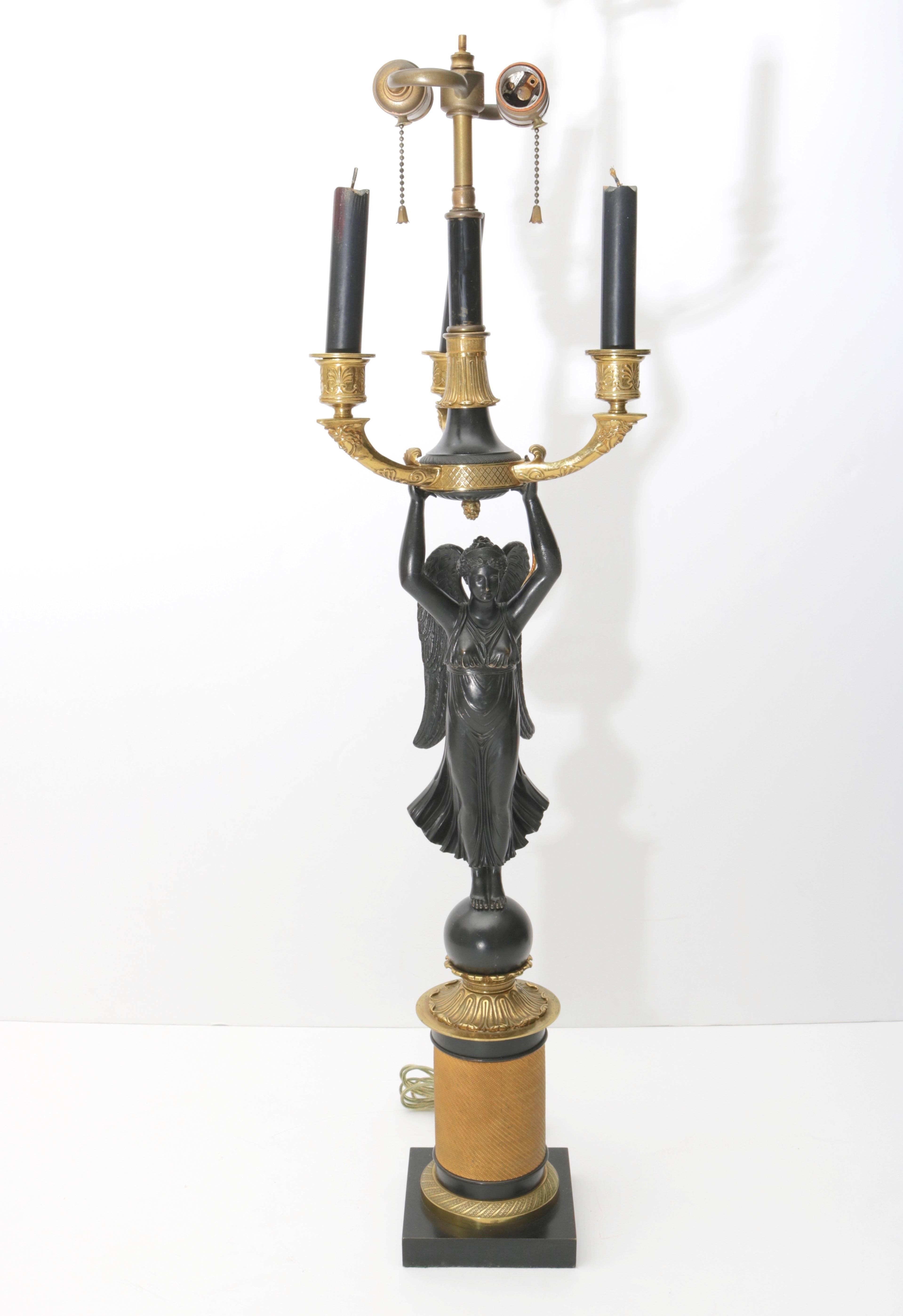 This stylish table lamp began its life as a candelabra and was converted to electric sometime in the 1940s. The piece is detailed with a central winged figure with upturned arms supporting three candle holders. The arms are detailed with the face of