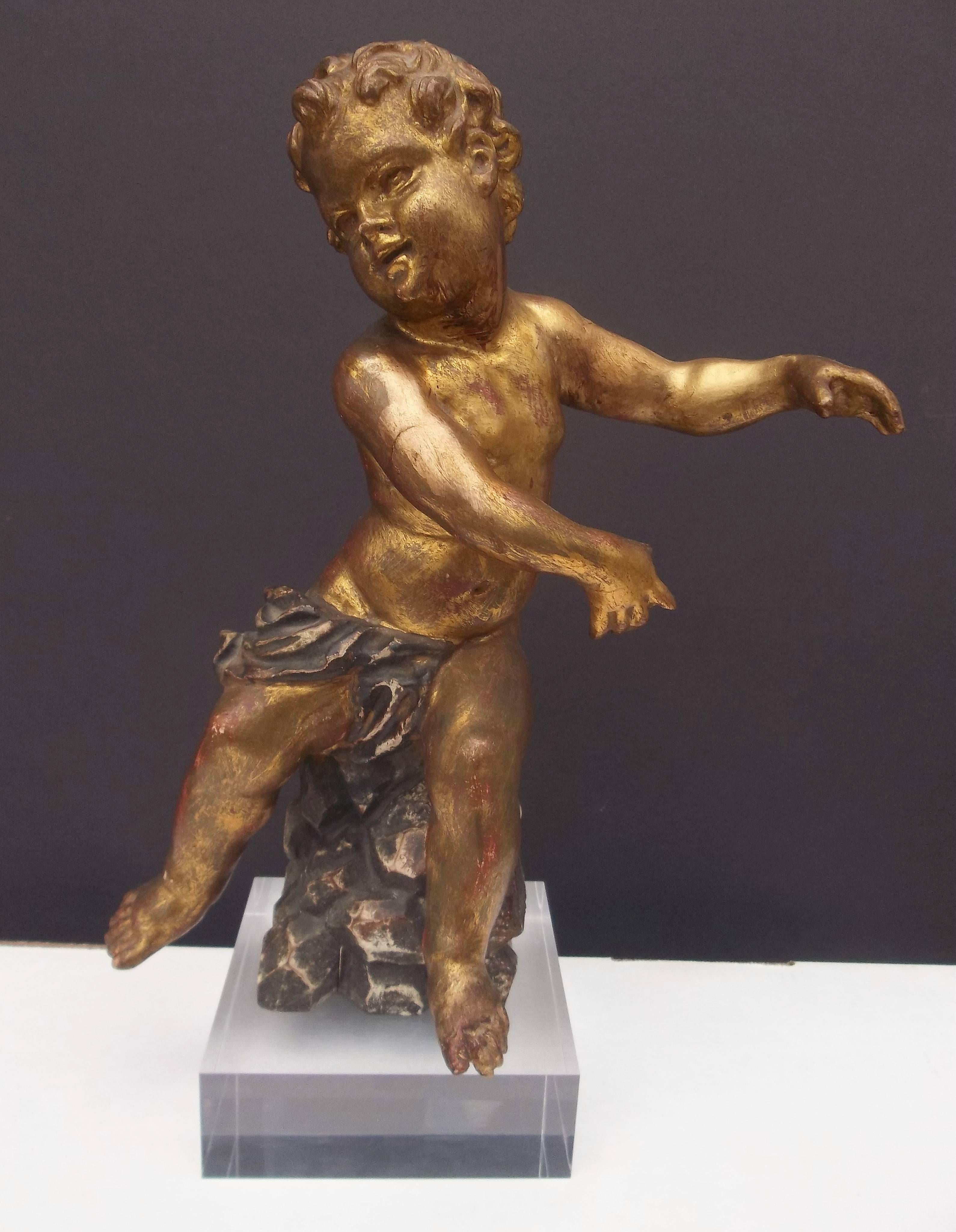 Cherub or putto, sitting on a faux rock and the whole on a later acrylic base . Very well detailed. Minor rubbing of the gilt to red bole. Joint shrinkage. Touchup to one toe. Wear to the drape and rock base. Highly decorative. Happy little