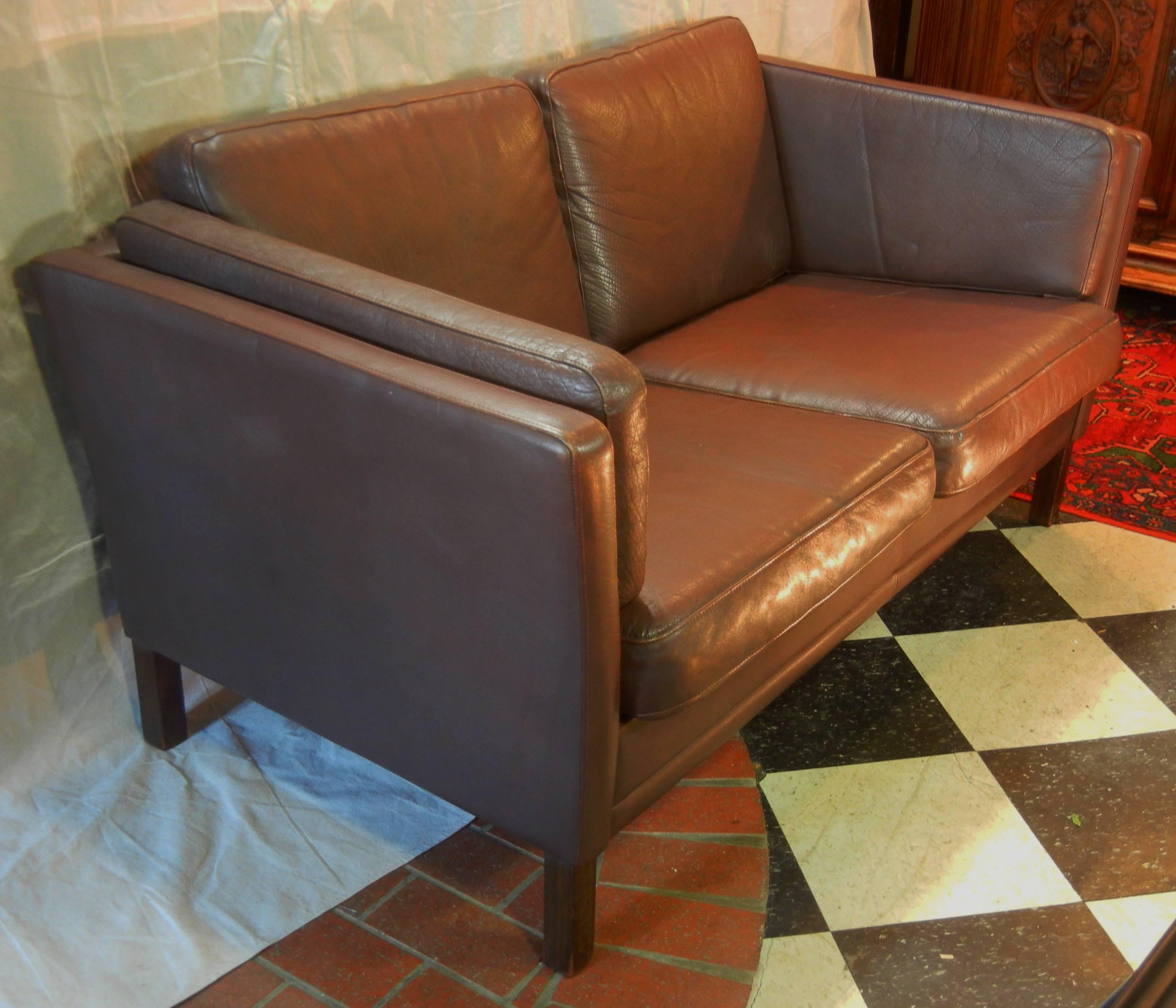 This loveseat or "two-seat sofa," is in the original chocolate brown leather upholstery, it has six loose cushions and it rests on square walnut legs. There is a slight outward lean to the sides which gives a comforting feeling of Good
