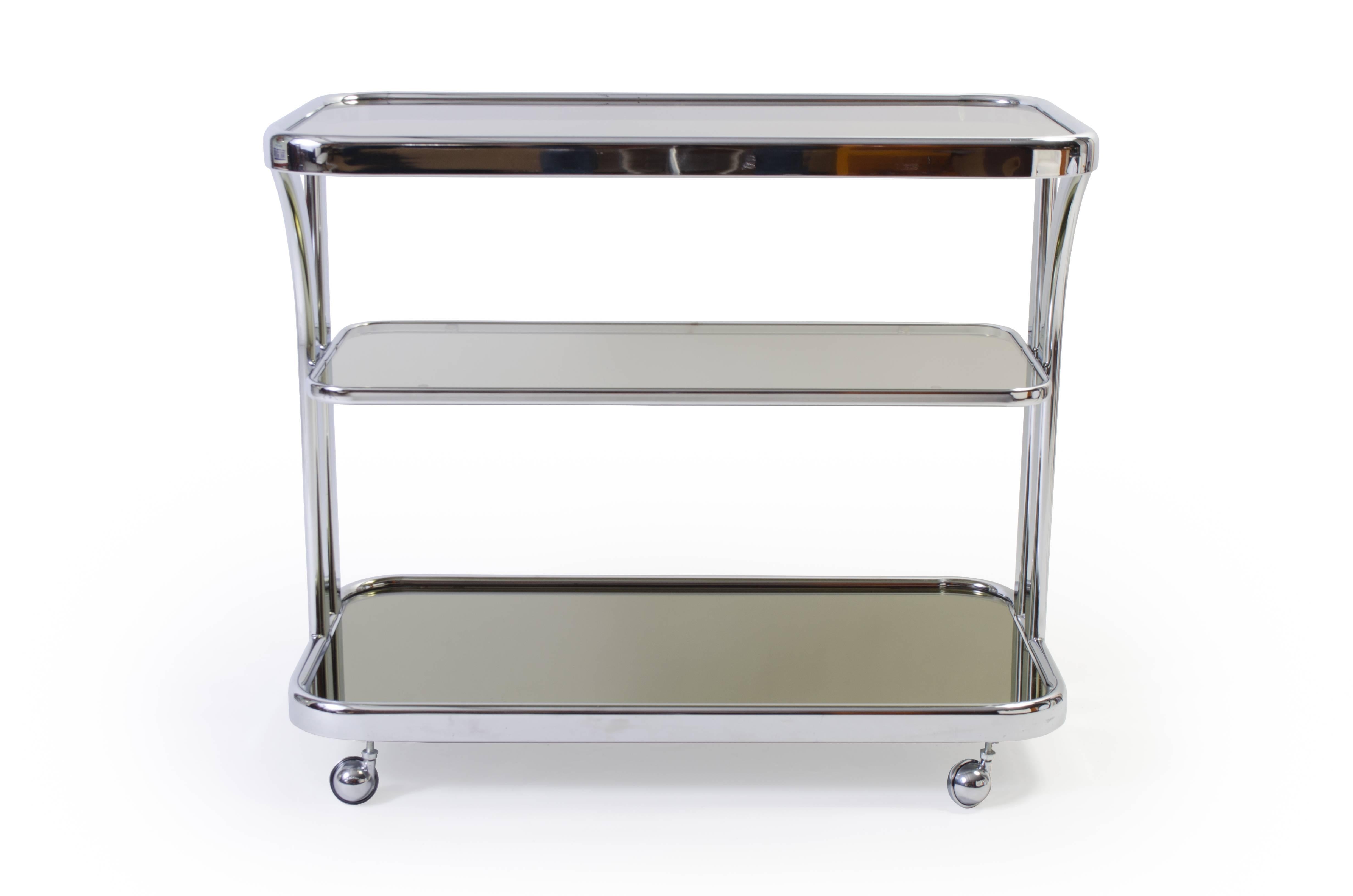A great design, for a great party. A stunning Art Deco motif bar cart featuring a chrome frame and three tiers. Each tier is surfaced with smoked glass with rounded corners fitting perfectly into the frame. Chrome ball casters finish this piece off,