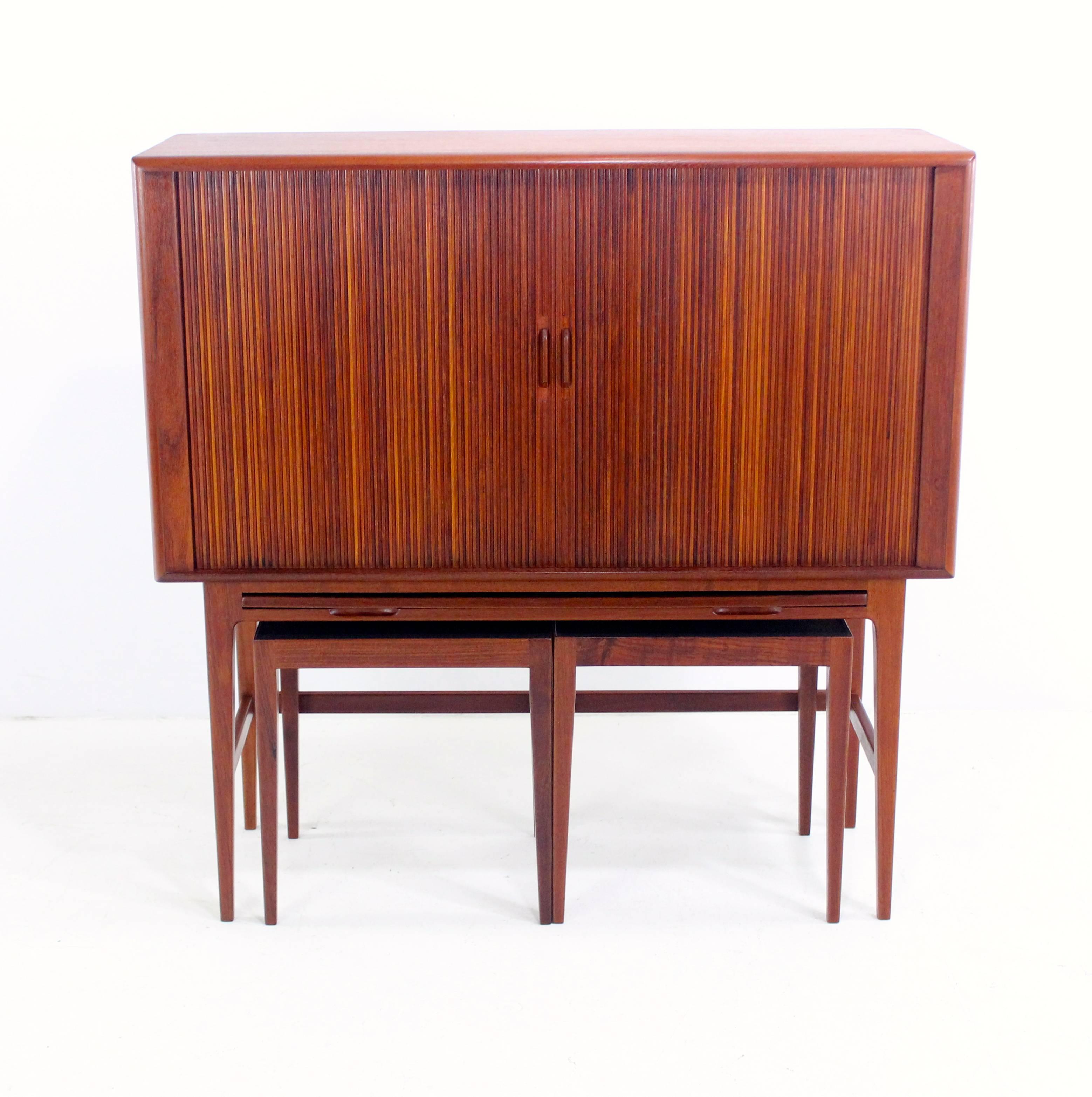 Danish modern bar cabinet with interior light designed by Kurt Ostervig.
Richly grained teak.
Tambour doors glide open to glass shelves and bottle storage.
Stain and heat resistant surface pulls out from cabinet bottom.
Two tables with stain and