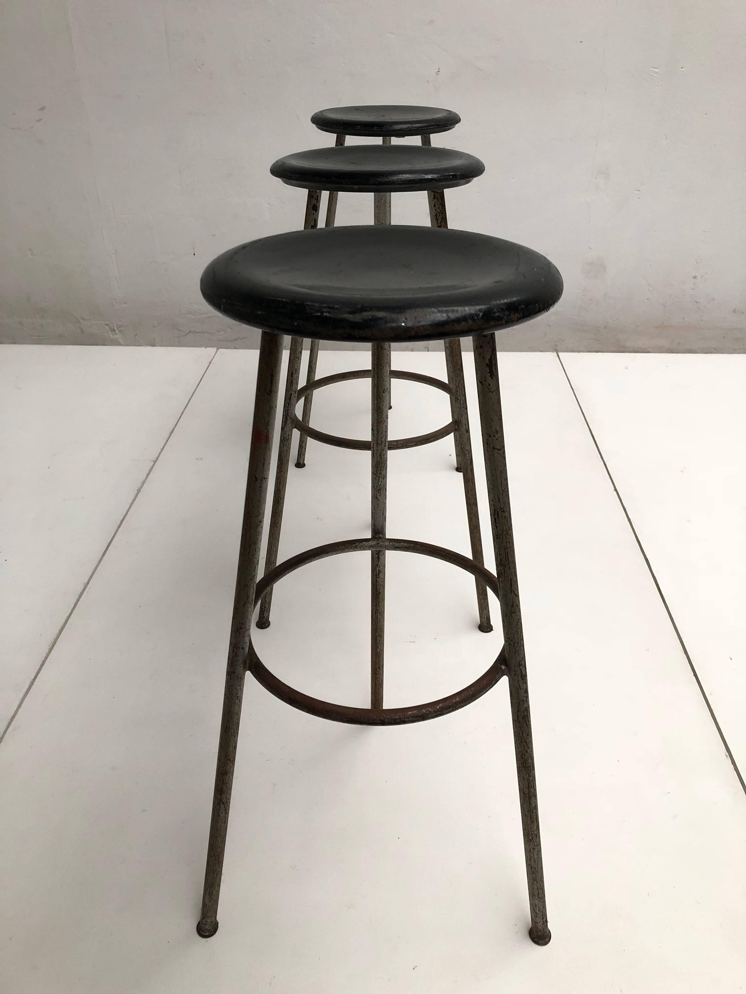 Unique set of three high tripod working / bar stools that originated from a Swiss confection Atelier 

Carved black lacquered wooden round seating on tripod tubular steel grey enamelled legs

All stools have a lovely old and aged patina from