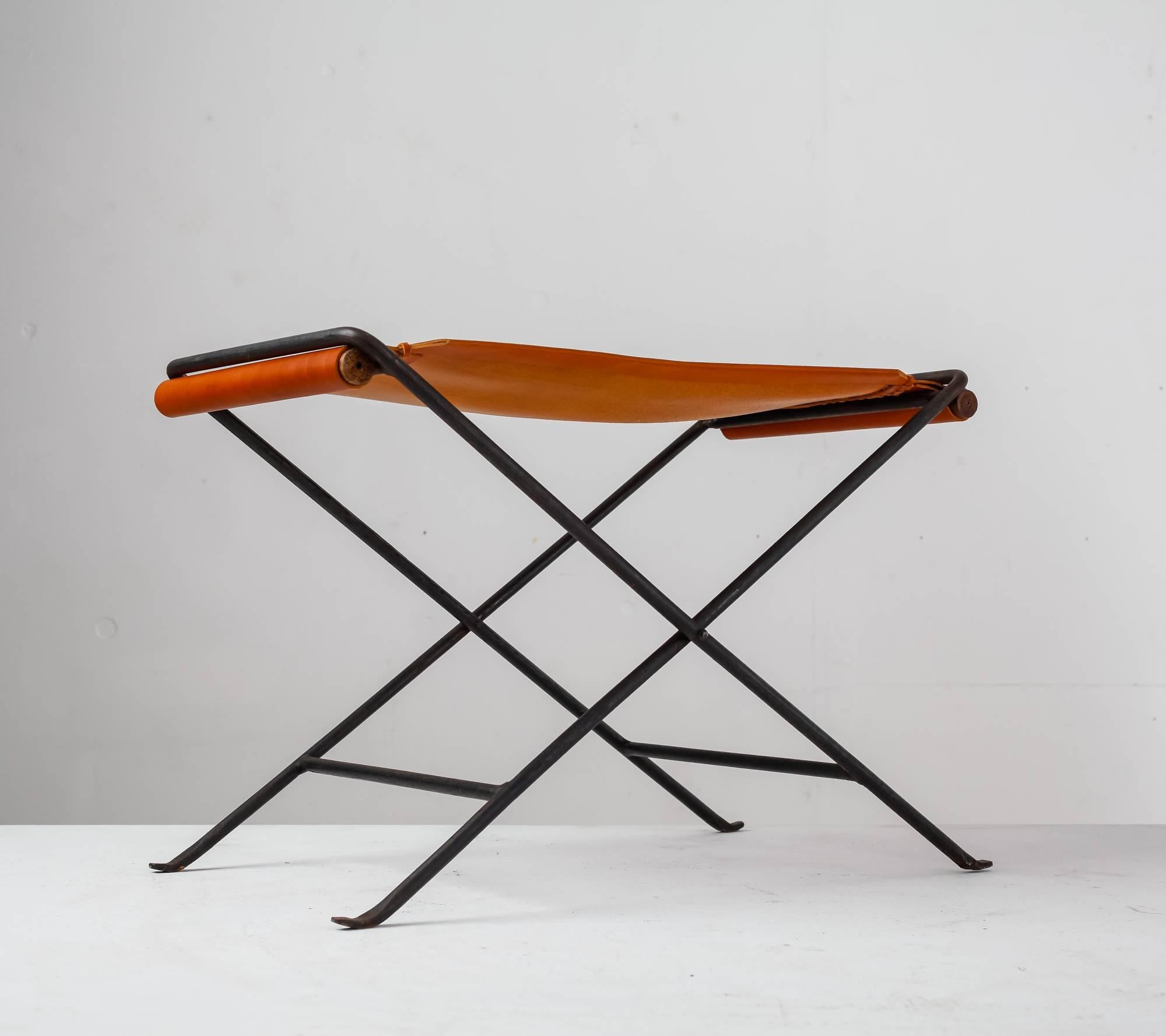 A foldable stool made of a black iron frame with a brown leather sling seat, attributeedto  Californian designer Cleo Baldon. The leather is fixed with two wooden poles. The stool has been professionally reupholstered in our in-house