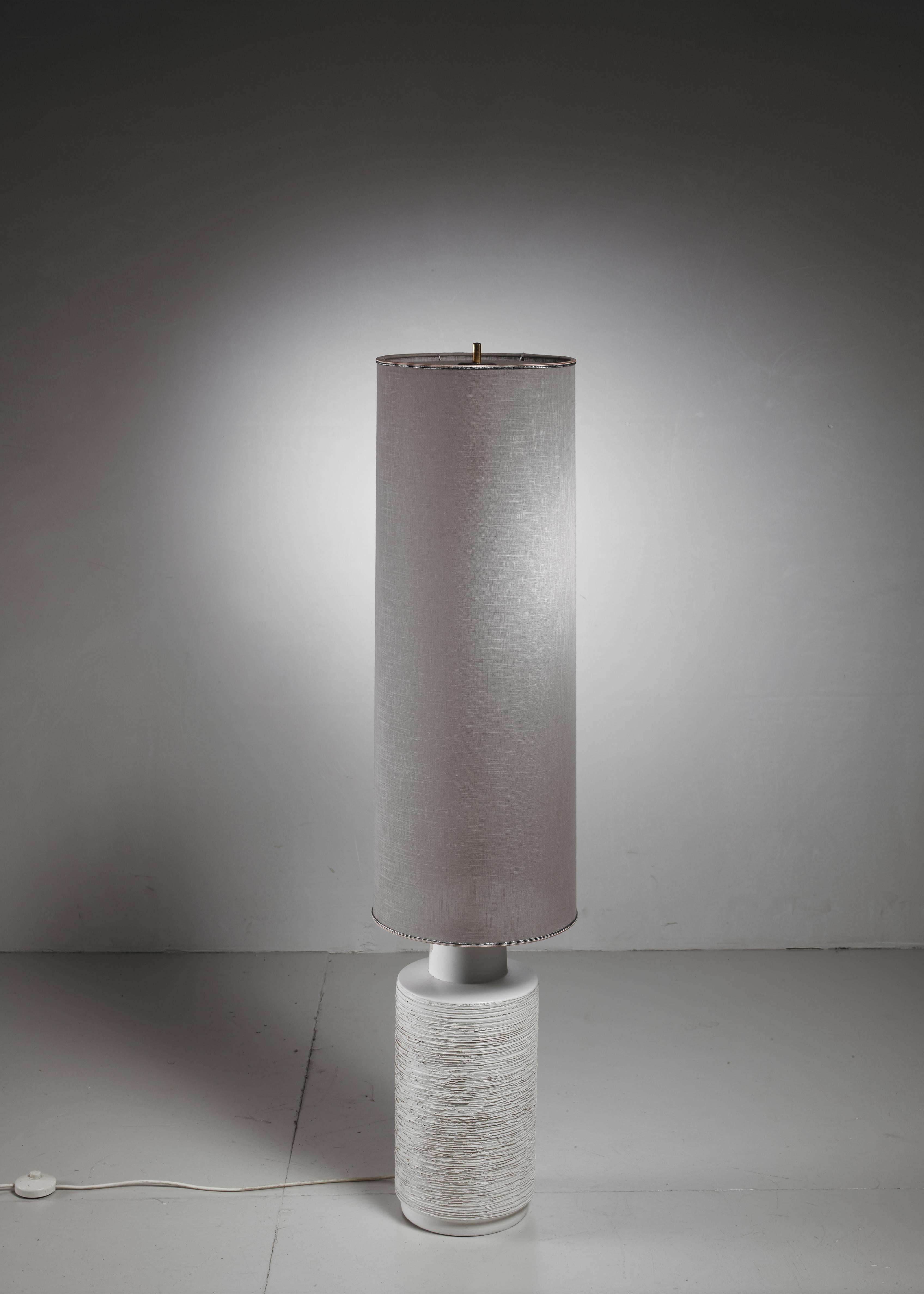 An off-white ceramic based floor lamp with a large cylindrical, grey linen shade.