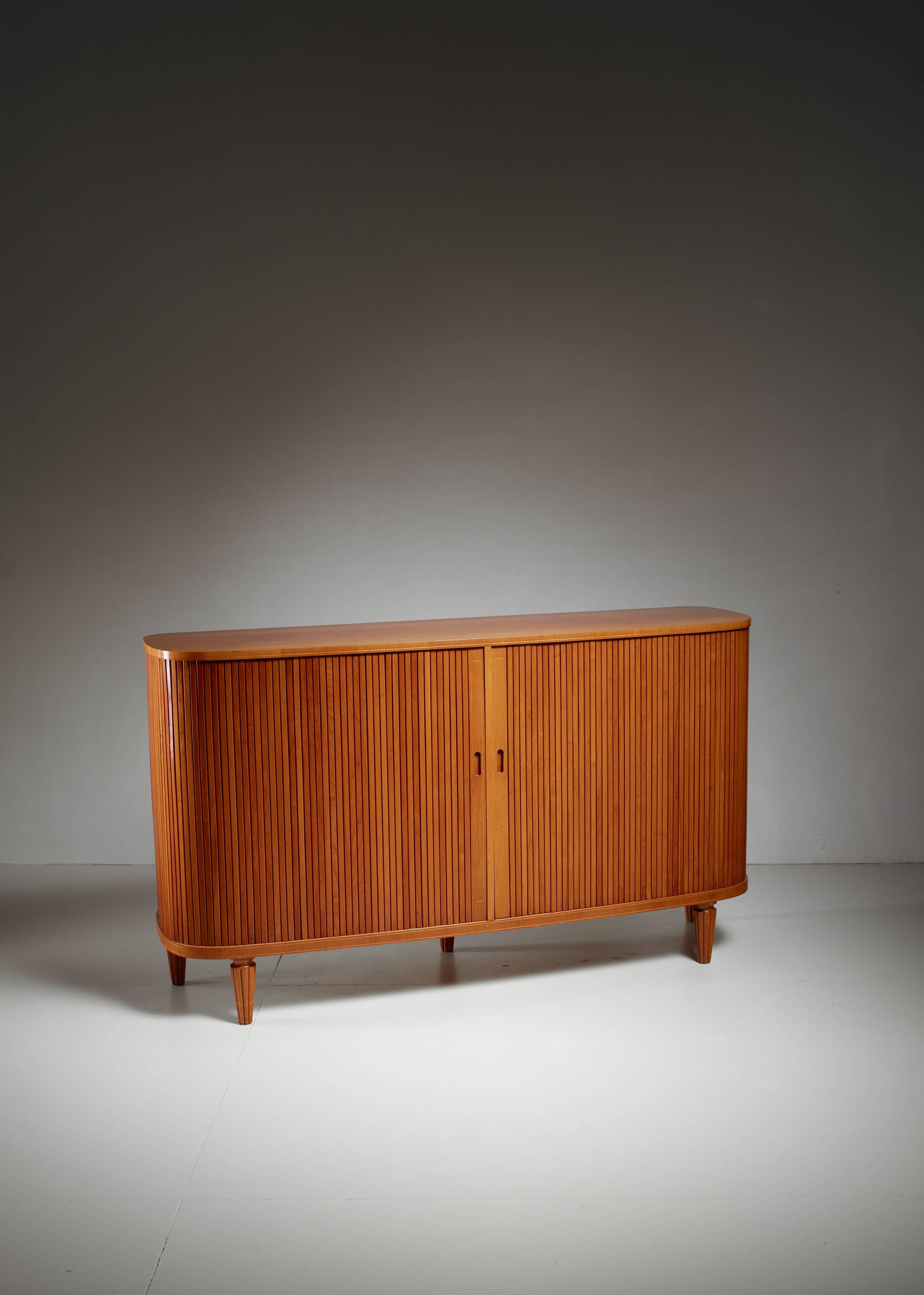 An elegant Swedish sideboard made of elm, with two tambour doors, standing on five sculpted, tapering legs. Inside the sideboard has shelves and five small document drawers. The edges underneath and above the door have been subtly decorated with