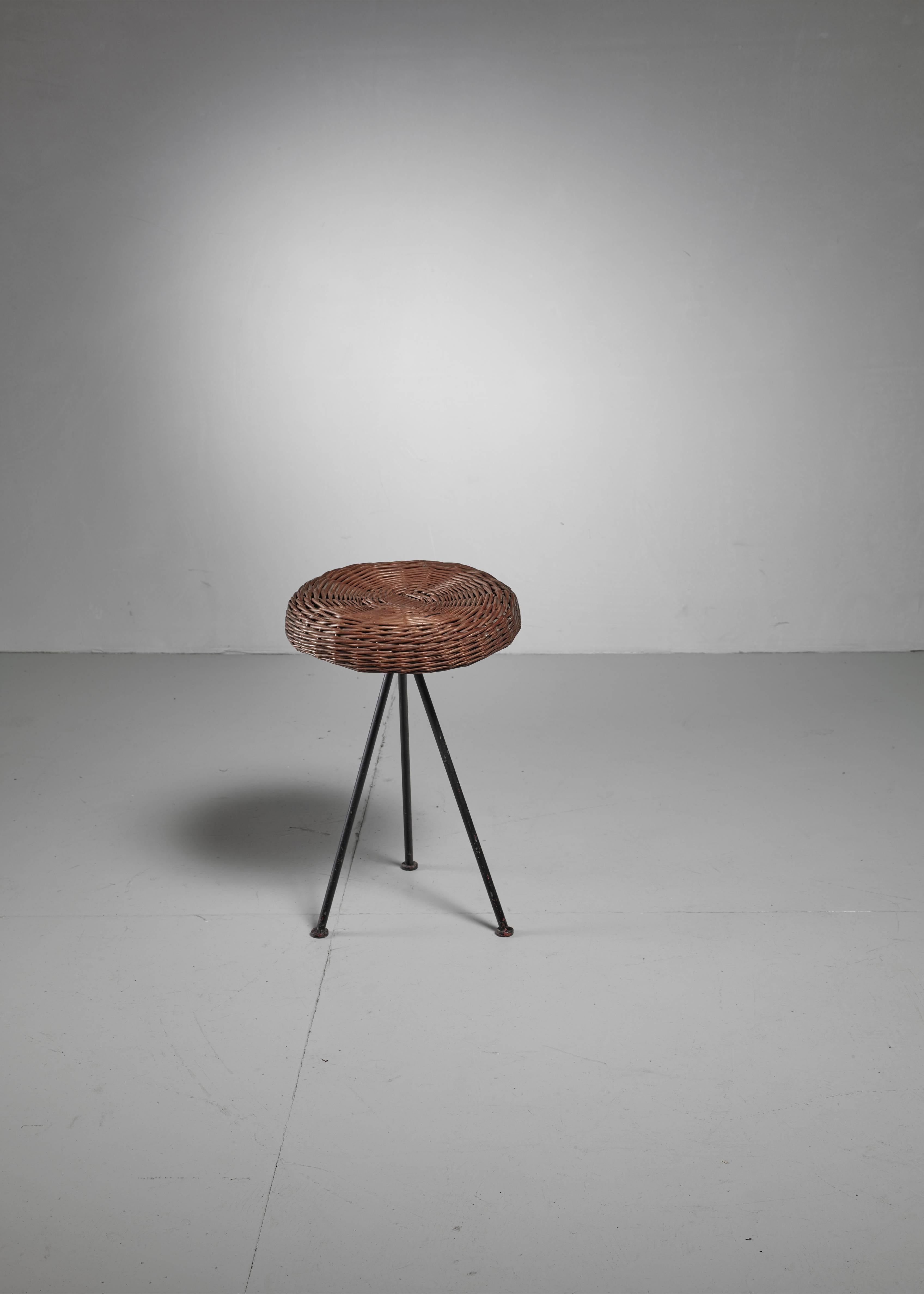 A 1950s American stool made of a black iron tripod frame with a woven rattan seating. The stool was designed by Norman Cherner for Konwiser.