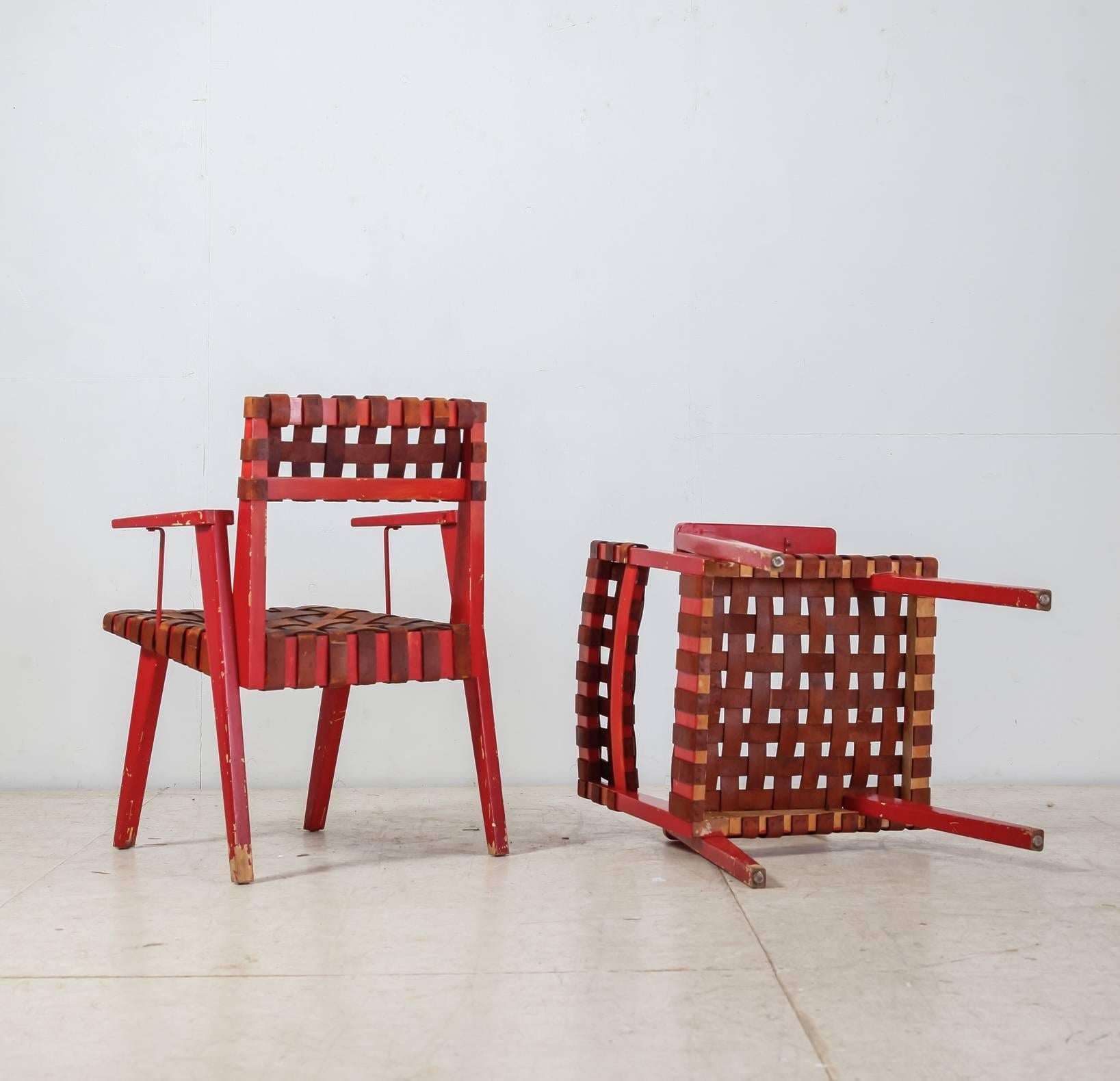 A pair of armchairs made of a red painted wooden frame with a brown leather webbing. The armrests are supported by a metal part.
The chairs are signed with 'Chairmaster Product' and 'Bronx, NY.'

There are damages to the paint, but the chairs are