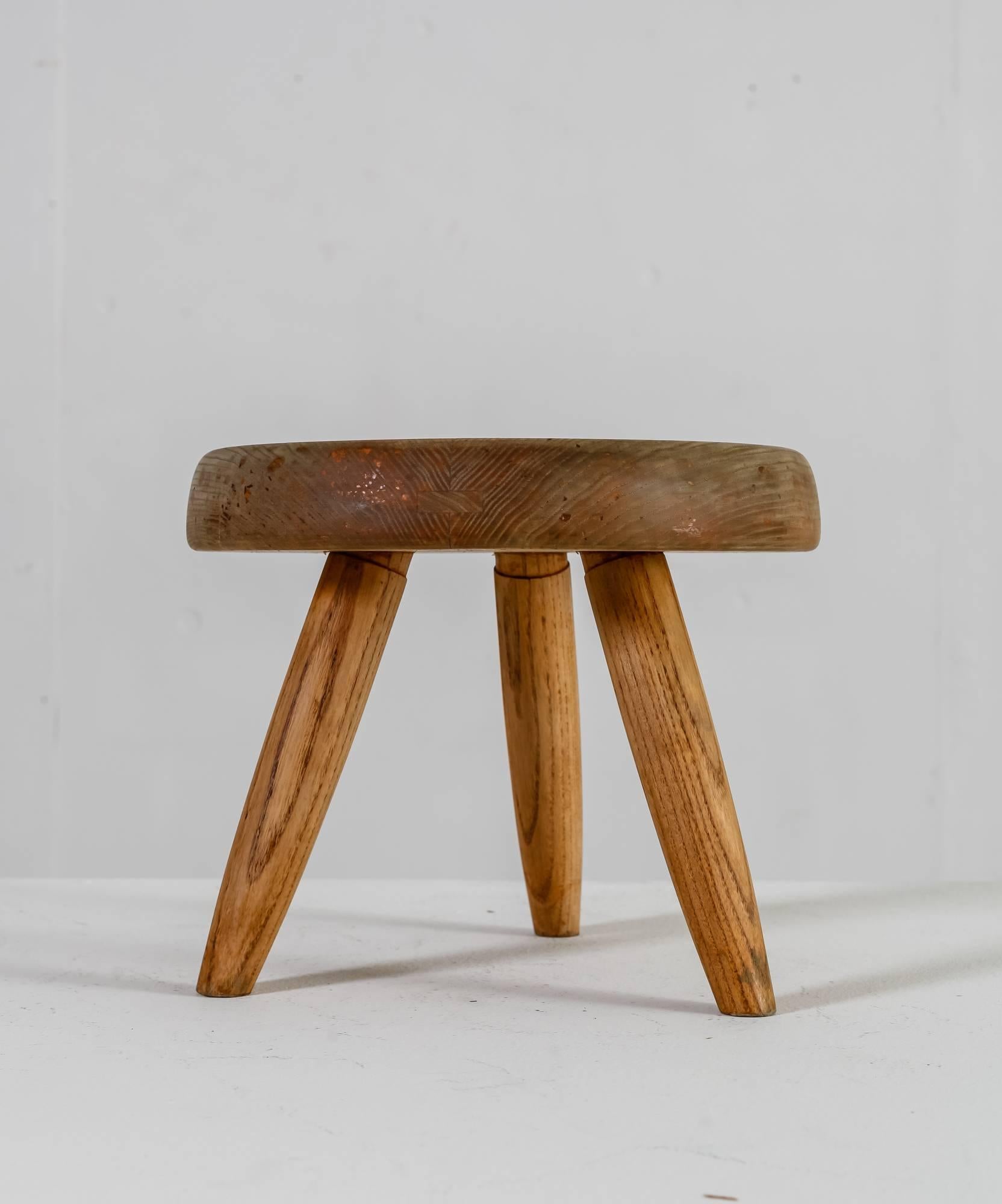 A low oak tripod stool with a thick round seating, by Charlotte Perriand. The wood has a stunning, heavy patina and beautiful wood connections.

The first tripod stool of this type by Perriand was designed in 1947 and produced by Georges