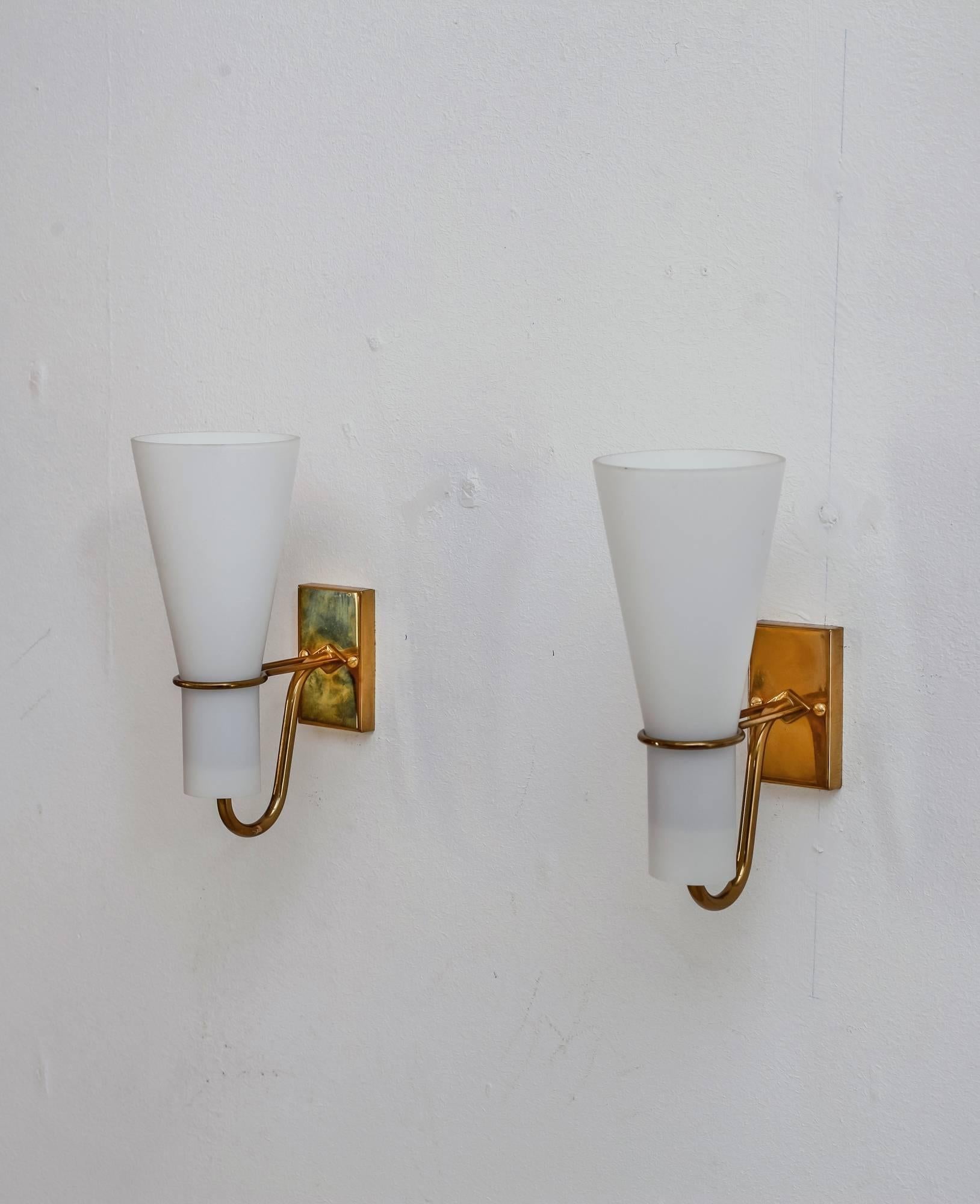 A pair of Asea brass and opaline glass bedside wall lamps. From the wall mount two arms extend that hold a trumpet shaped diffuser. 
One of the lamps is labeled by Asea and both lamps are in a perfect condition.