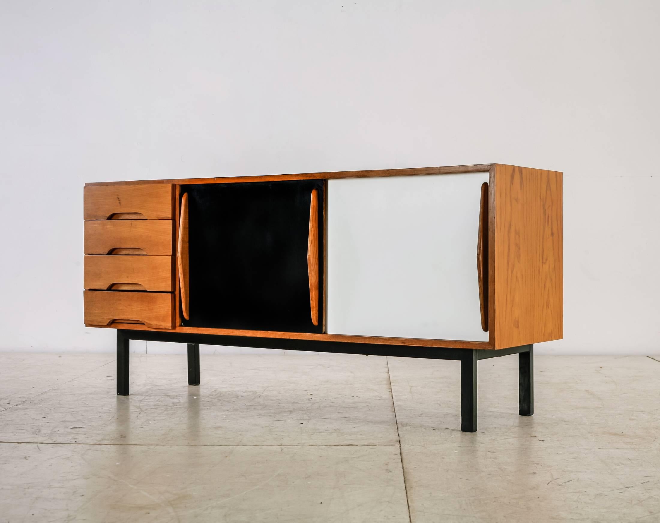 A Charlotte Perriand sideboard, made of ash with a black and a white formica sliding door. It has four drawers and stands on a black lacquered metal frame.
The sideboard was designed in 1958 for the Cansado new town in Mauritania and produced by