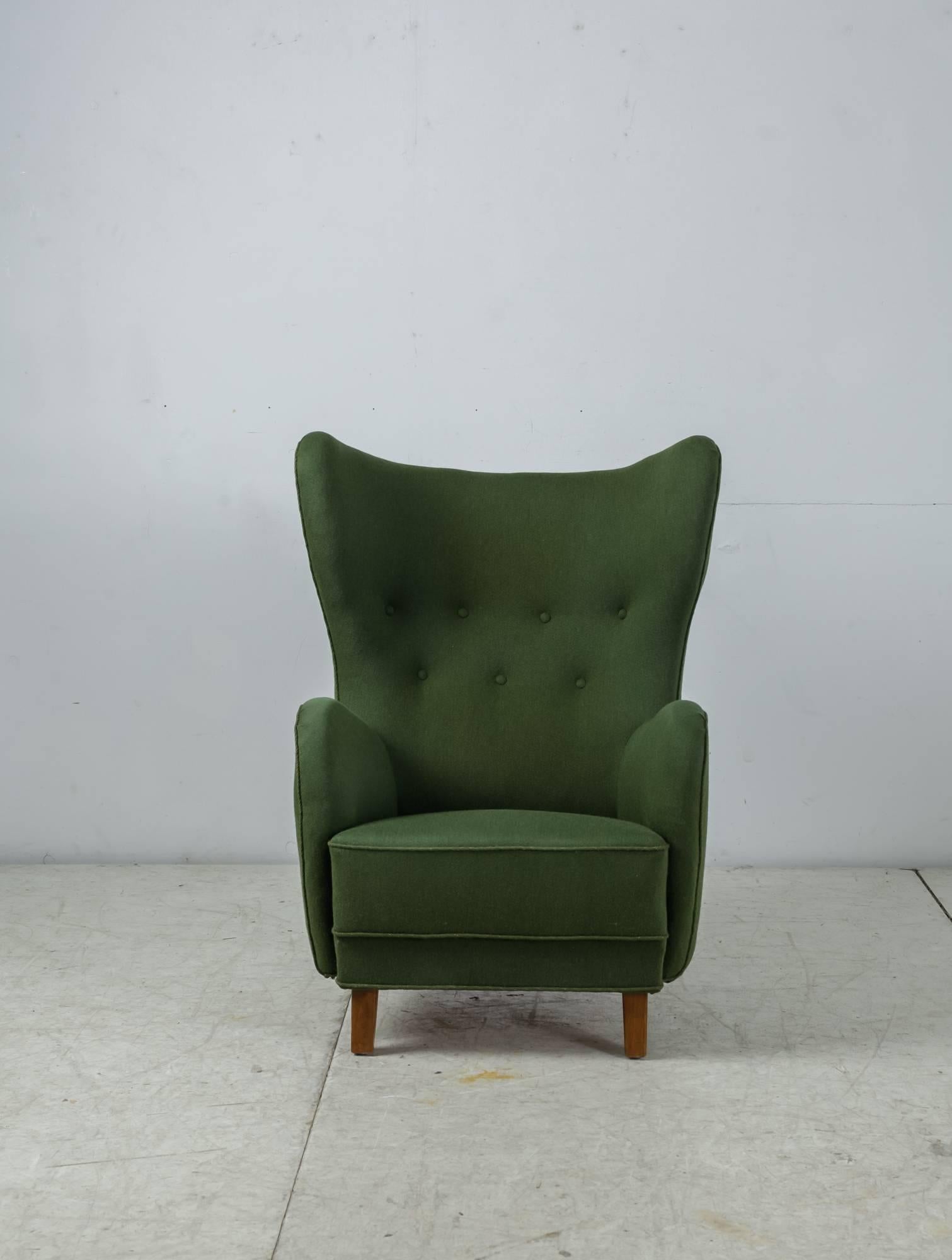 A high, wingback lounge chair in the manner of Mogens Lassen. The chair stands on stained beech legs and is upholstered with a green wool and the backrest is fitted with buttons.