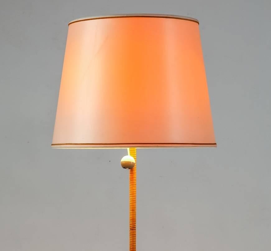 A brass floor lamp with a stem that has cane wrapped around it. The lamp has two light bulbs and a round off-white shade by Stockmann Orno.
The dimensions of the shade are 34.5 cm (13.5