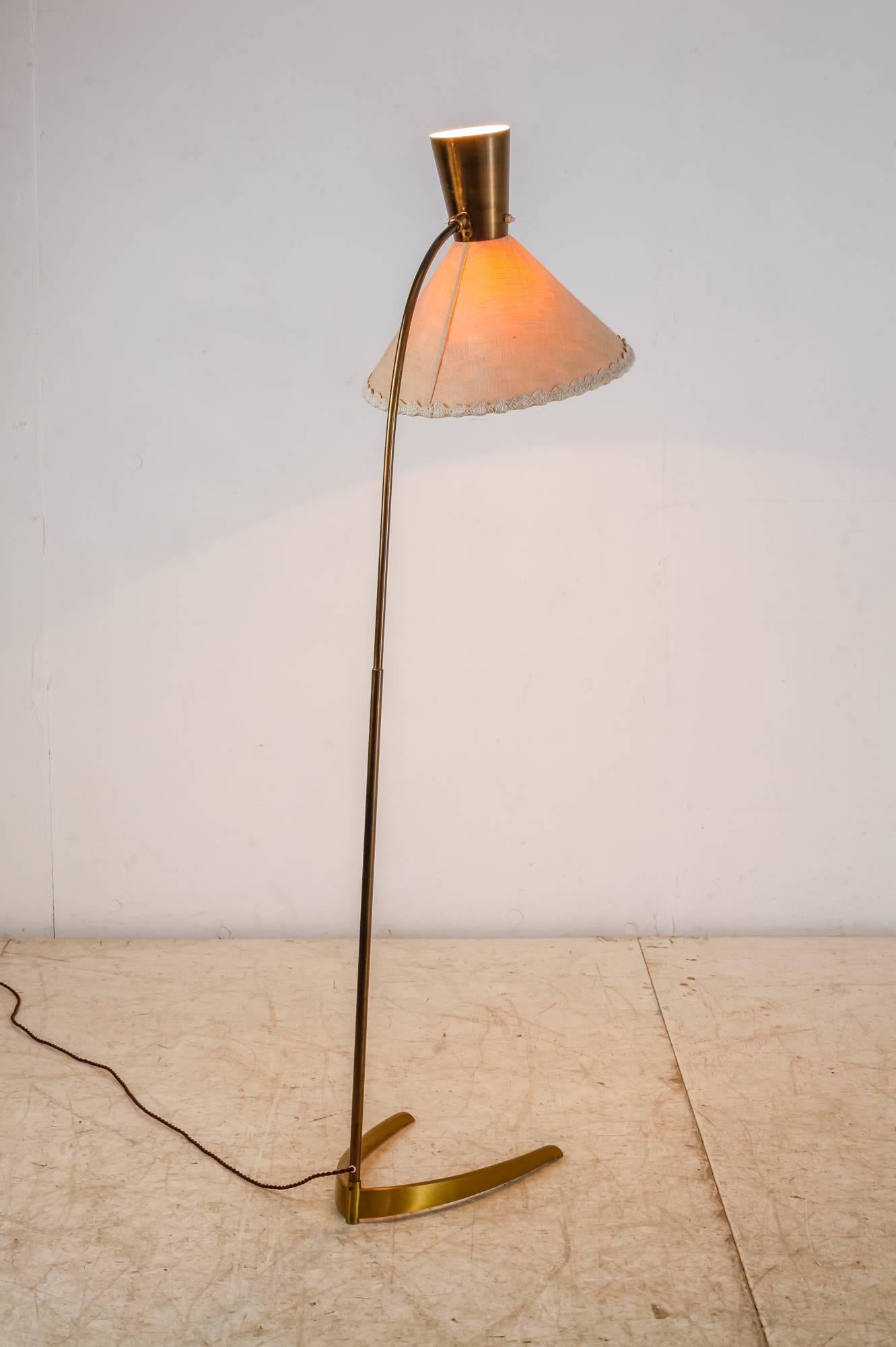 A 1950s brass floor lamp with a crow's foot and a diabolo shaped hood. The lower part of the adjustable hood is made of a beige fabric.
The lamp has two light bulbs, one uplighter and one shining down.
