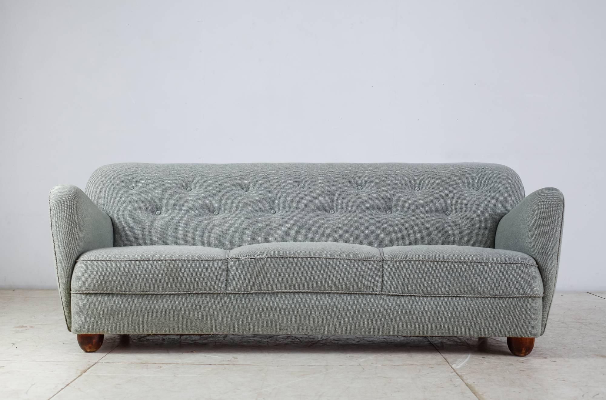 A slightly curved, three-seat sofa, standing on oak legs, round in the front and rectangular in the back. The sofa has a light blue fabric upholstery with a buttoned backrest.
Structurally the sofa is in an excellent condition, but the fabric shows