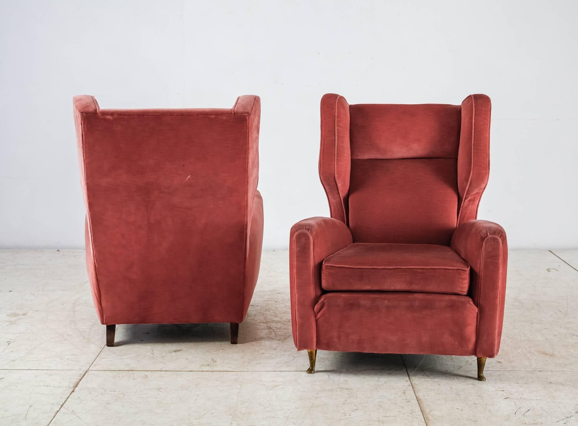 A pair of Paolo Buffa wingback lounge chairs with a coral red velour upholstery and a loose cushion. The chairs stand on curved brass front legs and wooden back legs.
The fabric shows wear due to usage, but the chairs are in an overall good