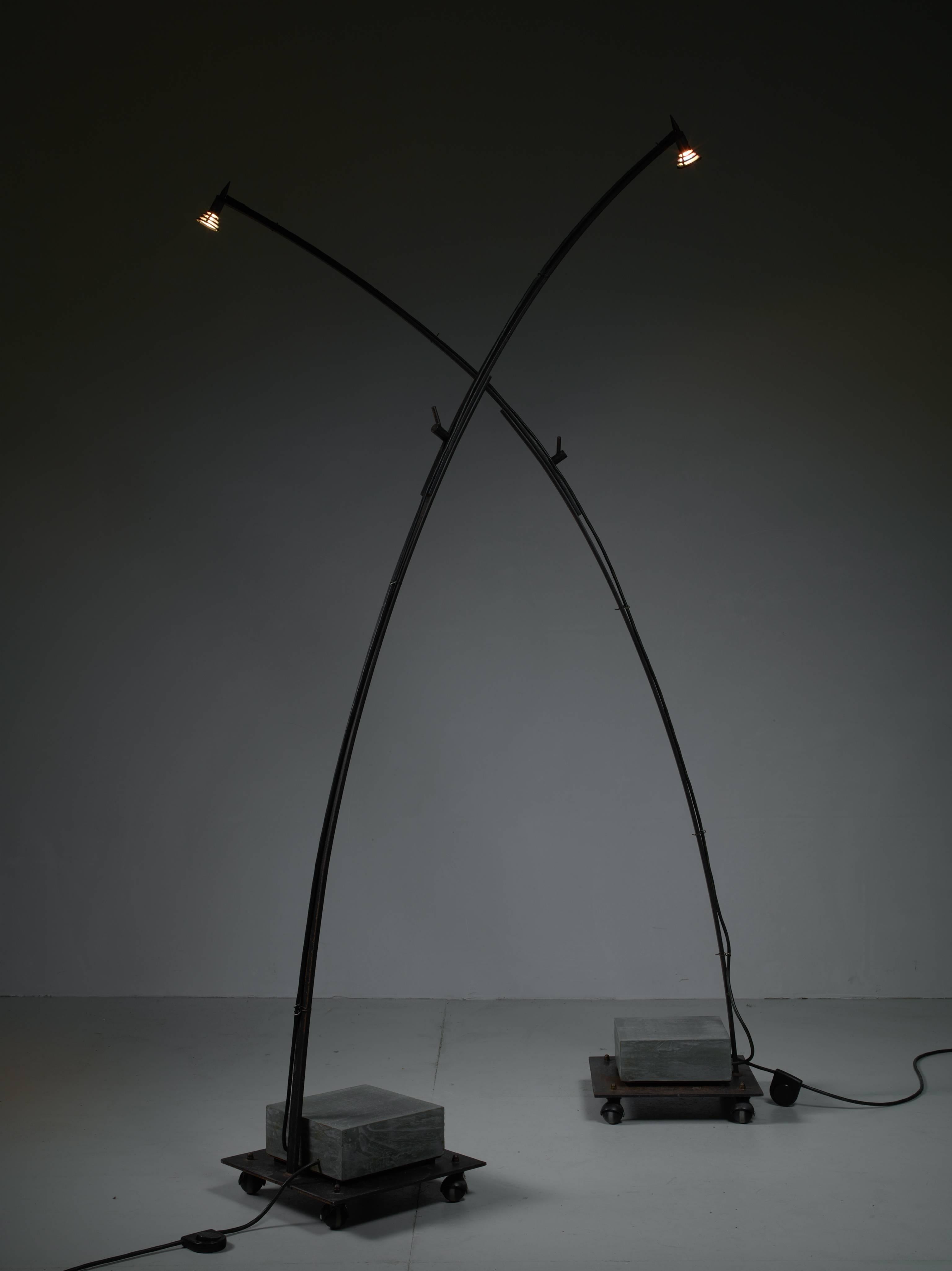 A pair of Industrial floor lamps made of metal.
They are reminiscent to the work of the Pentagon Group. 

The lamps stand on a square metal base with wheels and a concrete counterweight. The lamps have extendable arms with a halogen lamp bulb. When