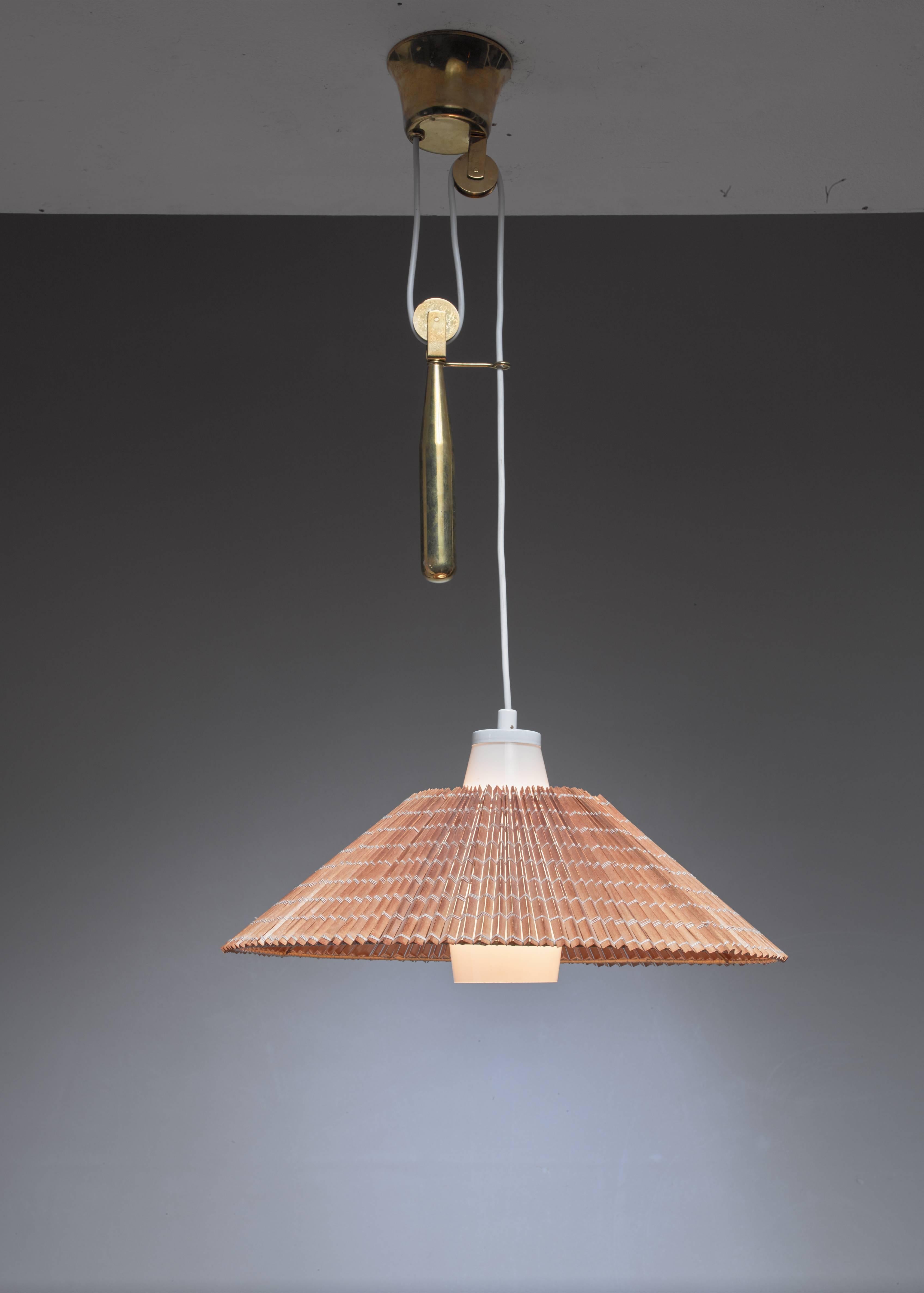 A Paavo Tynell pendant lamp with a heavy brass counterweight. The hood of the lamp is made of thin wood slats and rests on an opaline glass diffuser.