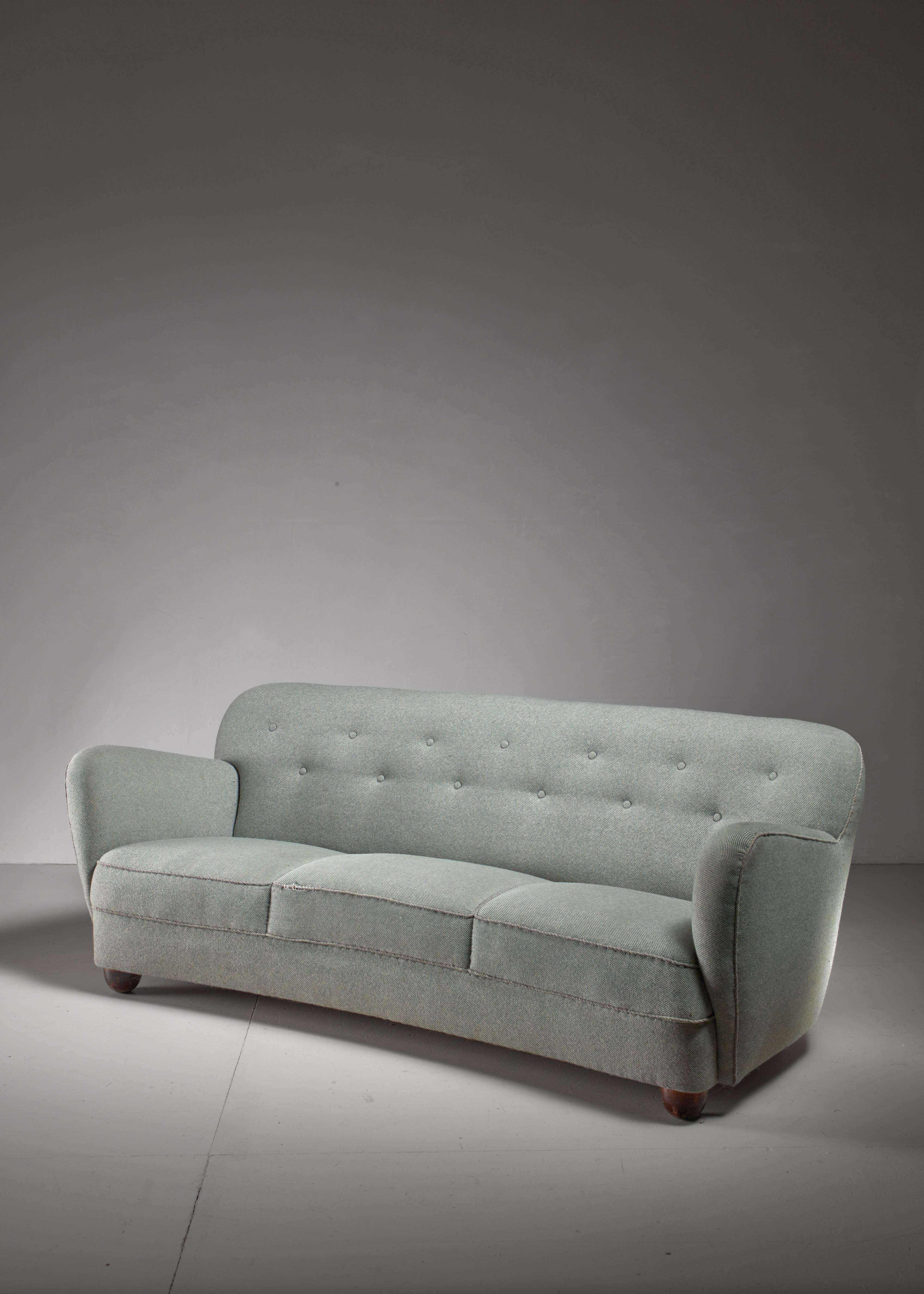 A slightly curved, three-seat sofa, standing on oak legs, round in the front and rectangular in the back. The sofa has a light blue fabric upholstery with a buttoned backrest. Structurally the sofa is in an excellent condition, but the fabric shows