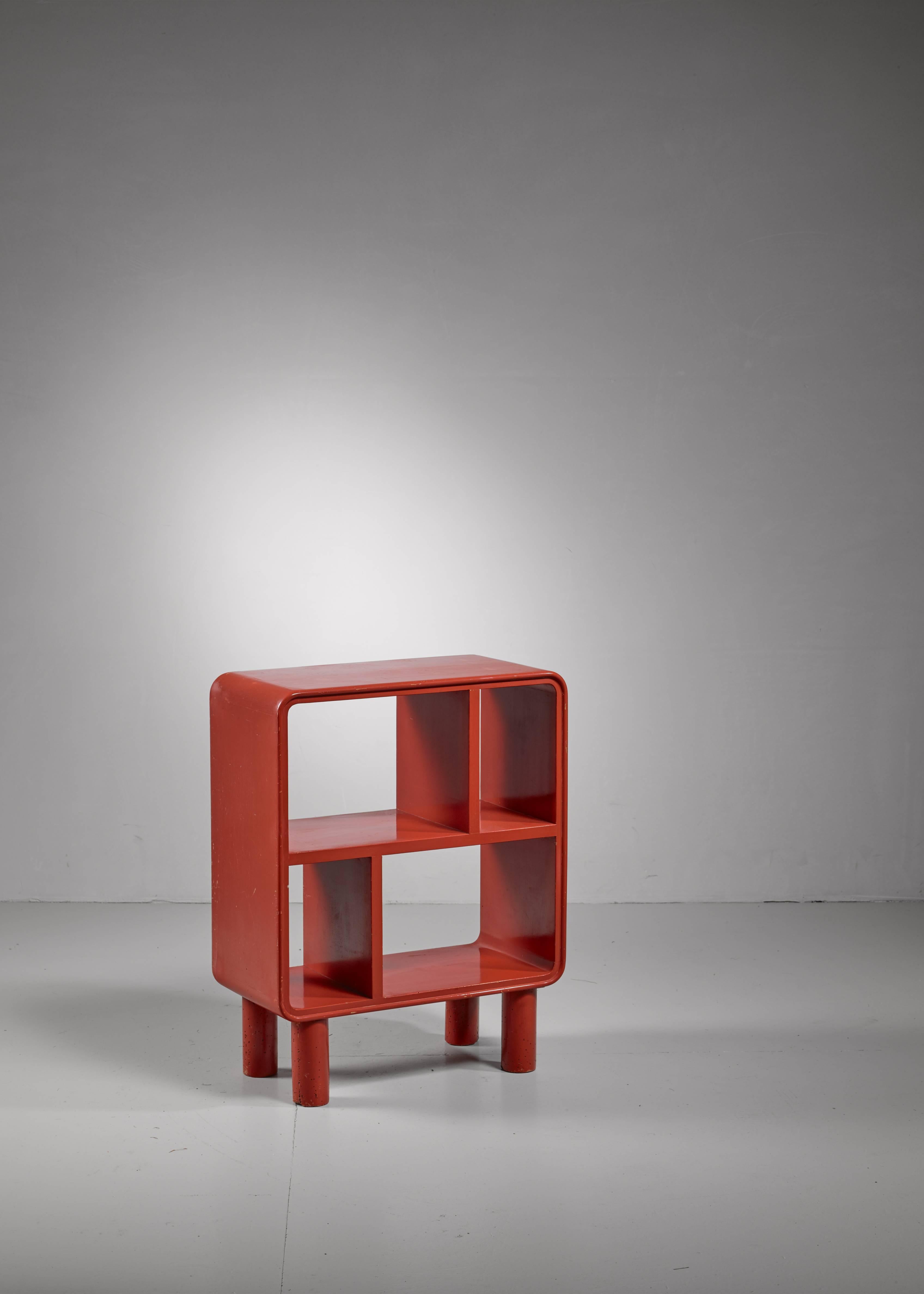 A small wooden shelving unit with four compartments, painted red. The square unit with rounded corners stands on four cylindrical feet.  