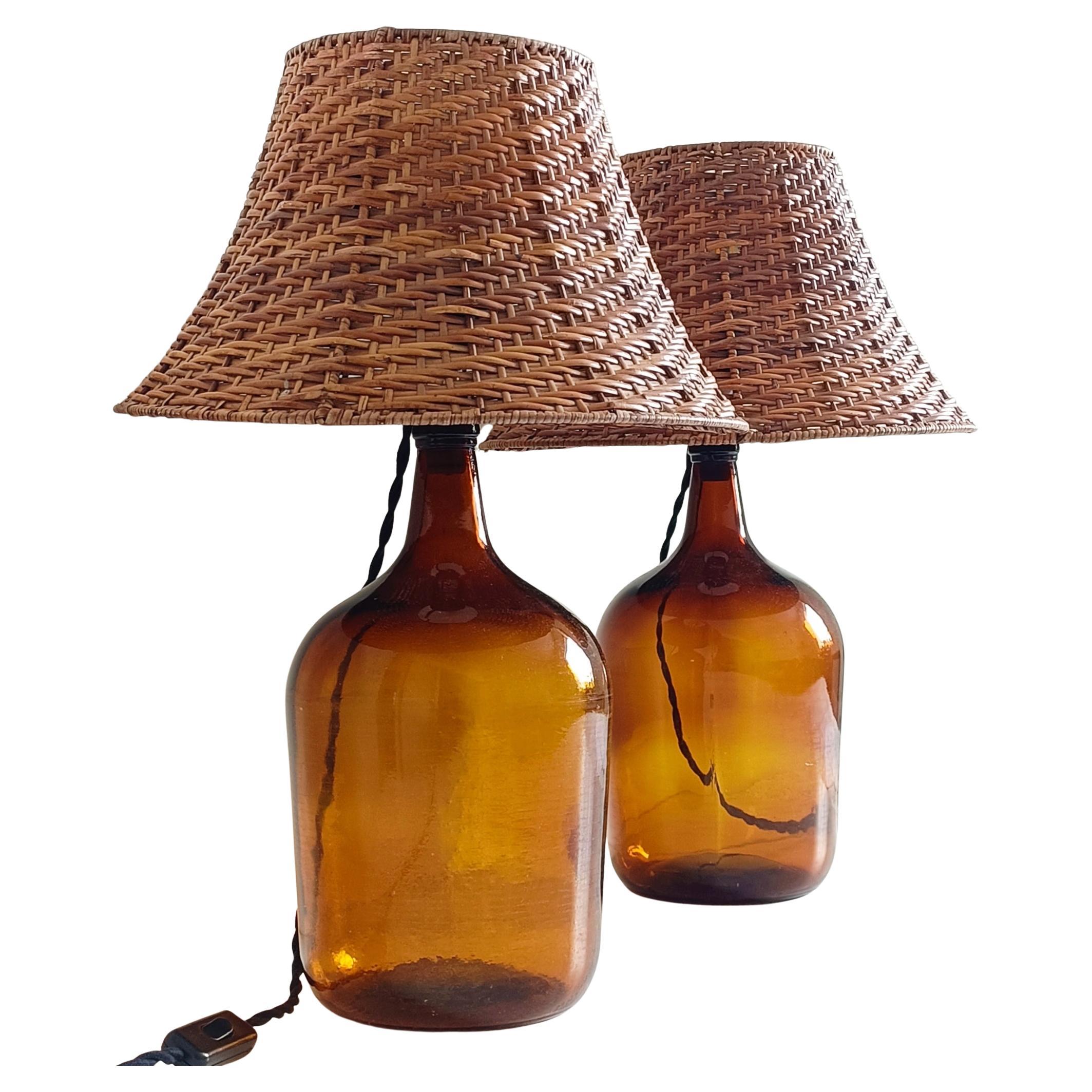 A gorgeous pair of table lamps made out of two demijohn amber color glass bottles and two Mid-Century Modern wicker lamp shades.

Demijohn glass bottles were used until the mid 20th century to sell wine in bulk in the old European wineries. 

This