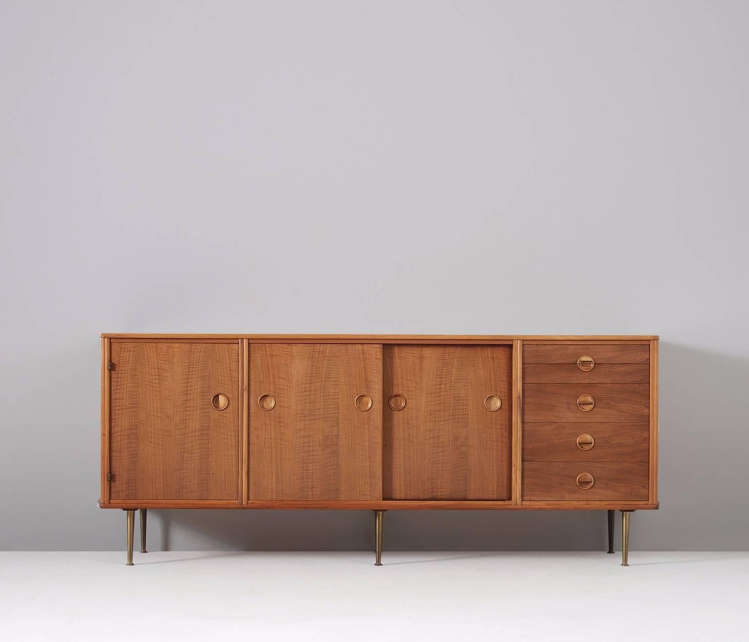 Sideboard in walnut and brass, by William Watting for Fristho, Netherlands 1958. 

Credenza in walnut with six brass legs. Five drawers on the right, two sliding doors in the middle and one door with shelves on the left. The watered walnut shows a
