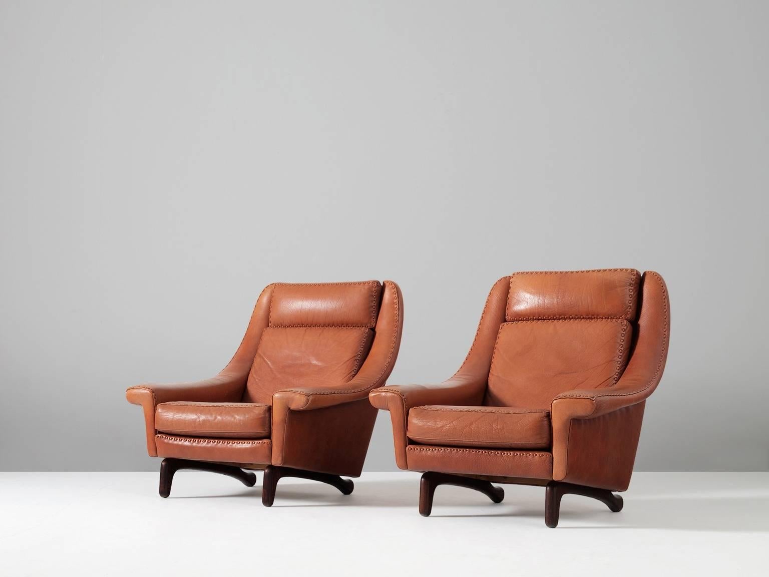 Pair of lounge chairs, in leather and wood, Scandinavia, 1960s. 

Highly comfortable lounge chairs in cognac leather. These wide chairs have a real interesting wooden base, with two legs. The dark stained wood has an anthropomorphic or organic