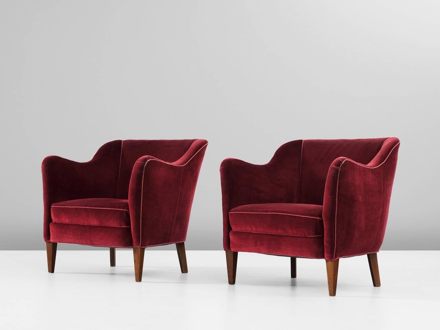 Pair of armchairs, in fabric and beech, Scandinavia, 1960s. 

Elegant pair of lounge chairs in red velvet upholstery. These chairs show some nice curves. The tight outside makes a nice contrast to the more soft inside. The armrest smoothly run