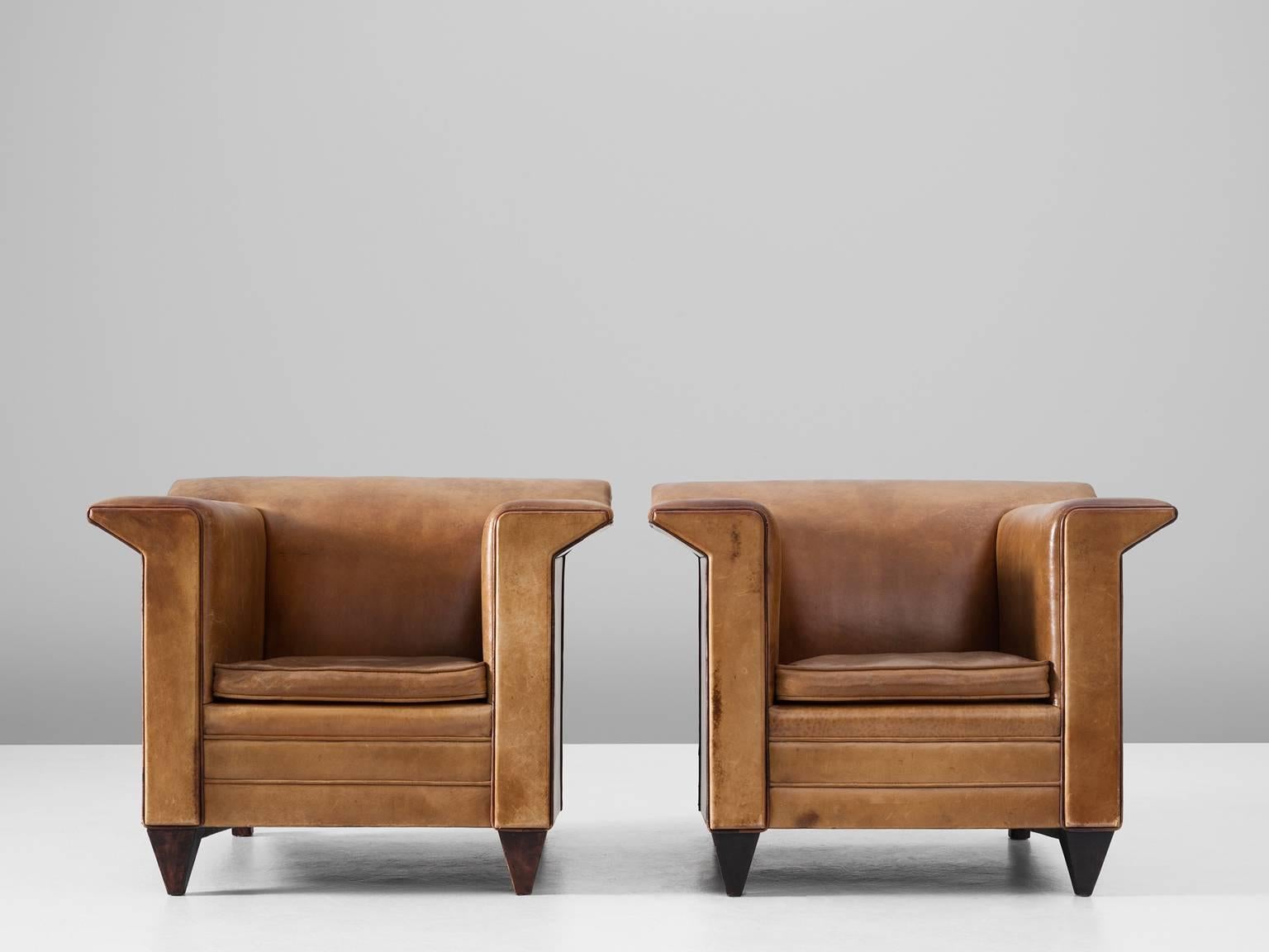 Pair of armchairs, in leather and wood, Europe, 1960s. 

A pair of cubistic clubchairs in cognac leather. The sturdy design has an interesting base that gives the model a certain lightness. The masculine shape of these comfortable chairs provides
