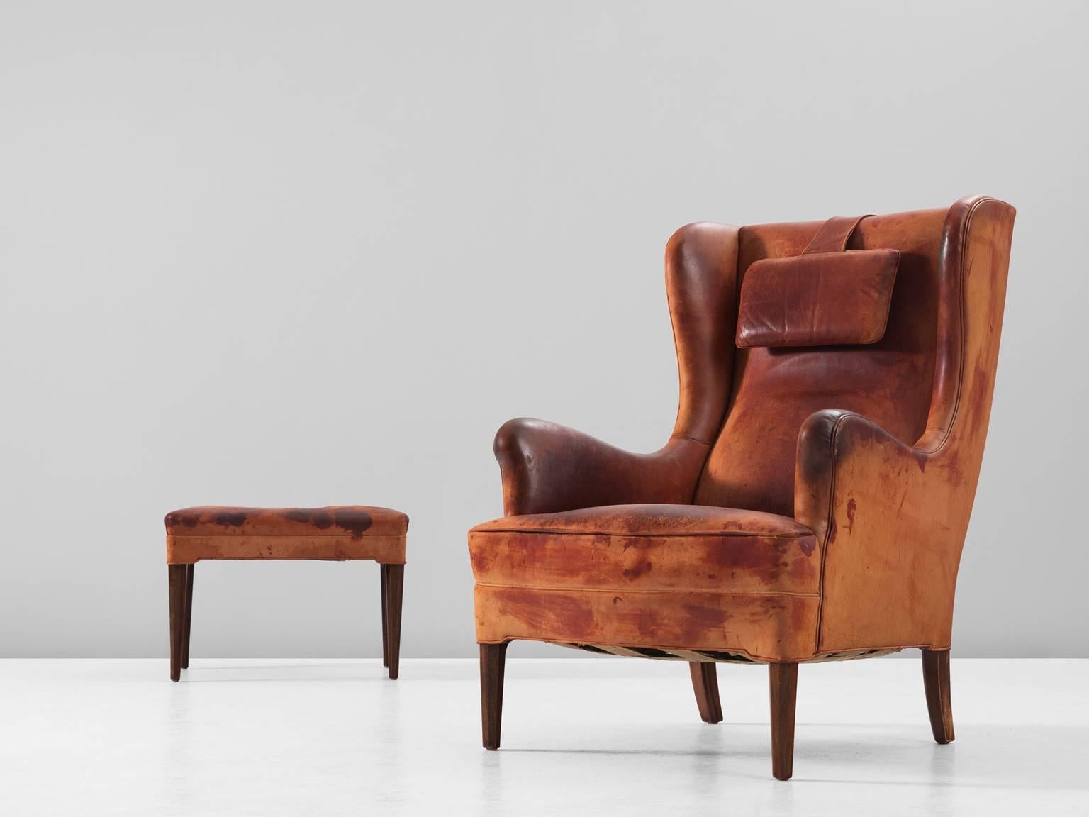 Wingback chair and ottoman in leather and wood by Frits Henningsen, Denmark, 1940s.

Classical wingback chair with accompanying footstool in patinated cognac leather. This chair and ottoman hold their original cognac leather upholstery. Therefore