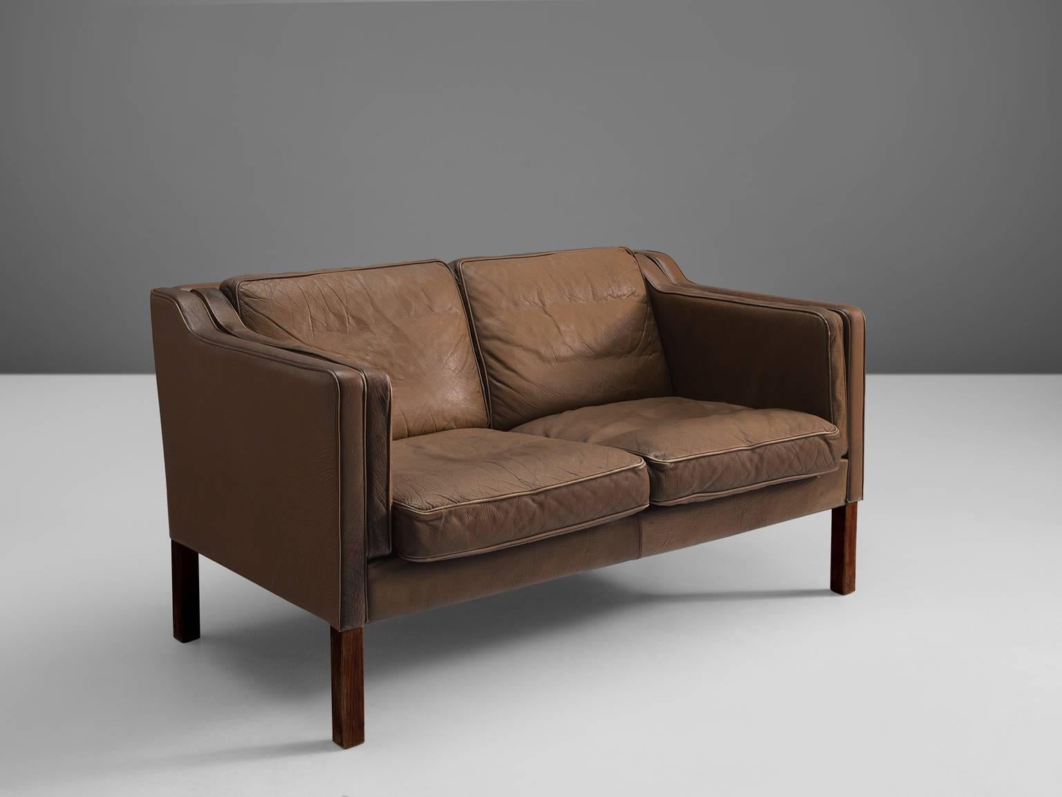 Two-seat sofa for Stouby Polster Møbelfabrik, leather and oak, Denmark, ca. 1960. 

Very well conditioned two-seat sofa. The brown leather shows a beautiful patina. Seating and back with dawn filled cushions. The wooden high legs provide a more open