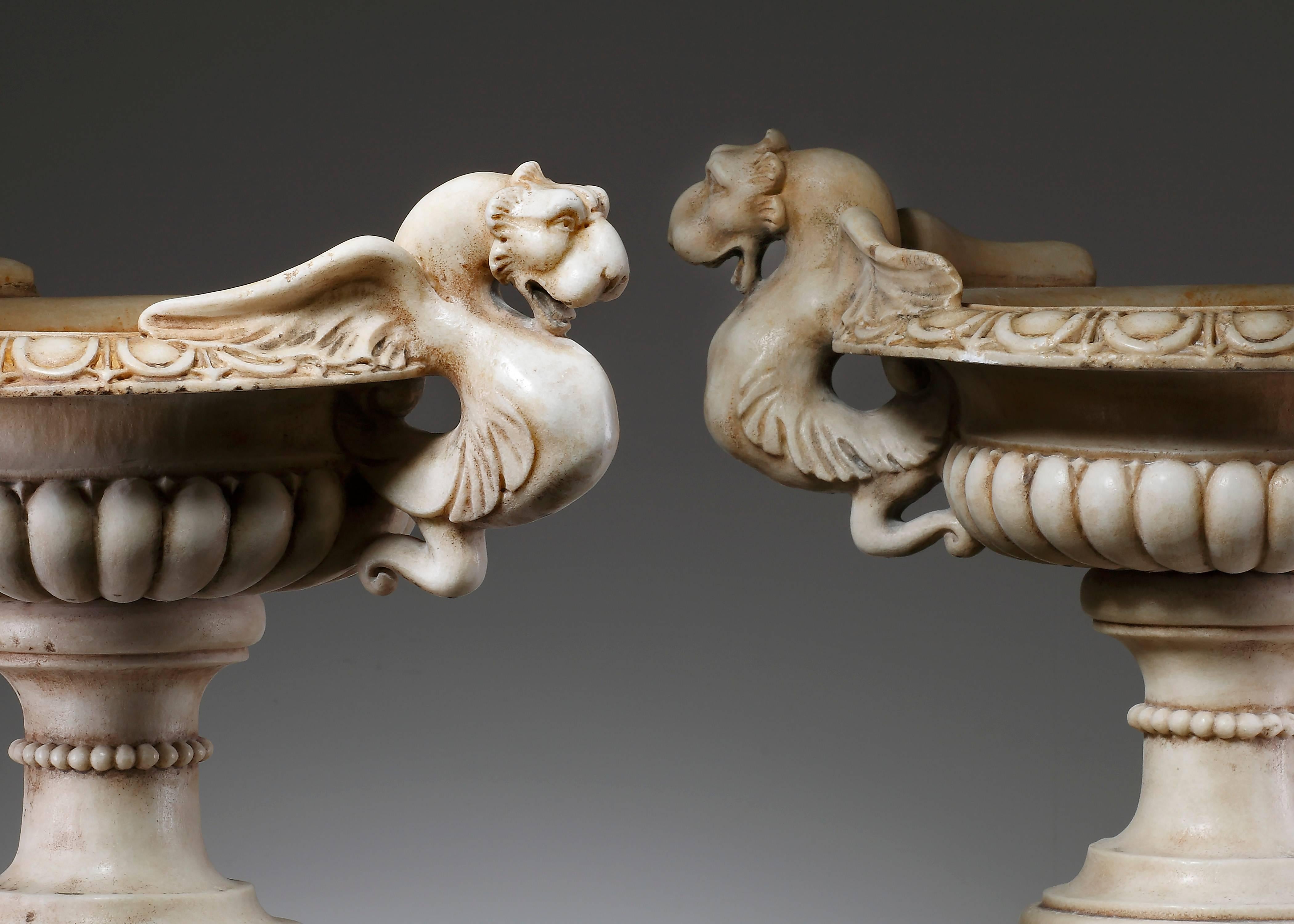 A pair of continental sculpted white marble garden urns, 20th century. Decorated with carved egg-and-dart rim and twin winged zoomorphic handles. Beaded stems and square socles.
