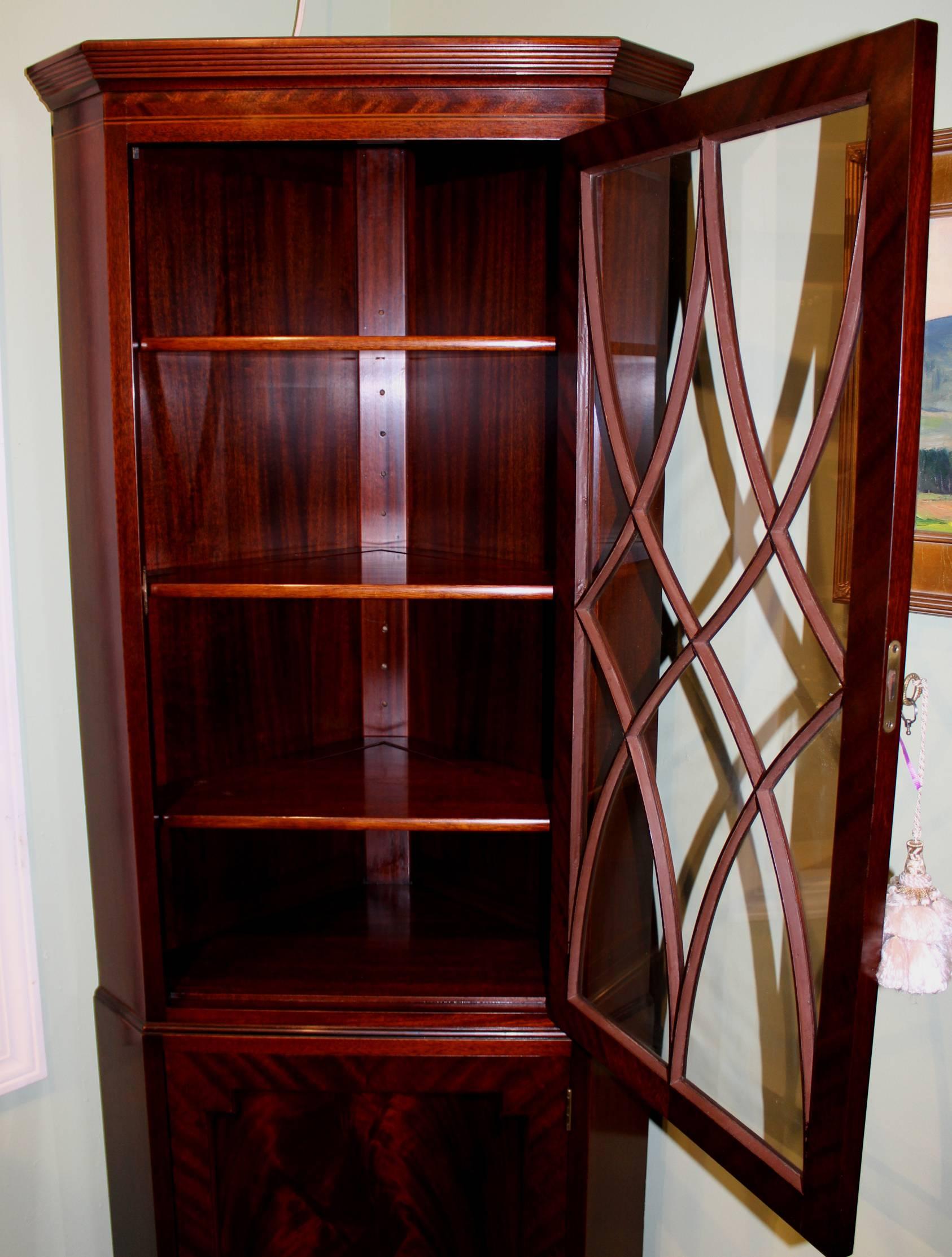 A splendid pair of mahogany corner cabinets made by Old Colony Furniture of Boston MA, in the English Sheraton style. Each with glazed upper door opening to reveal interior shelves. The base cabinet door with crotched mahogany veneer inset paneled