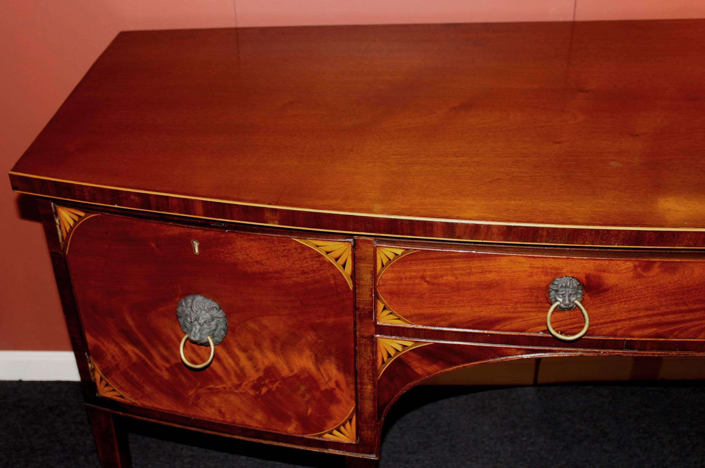 Georgian bowfront sideboard in mahogany with corner fan satinwood inlay, lion head ringed pulls, a central frieze drawer with loose felted flatware insert, flanked on the left with a single door opening to a one shelf interior compartment, and on