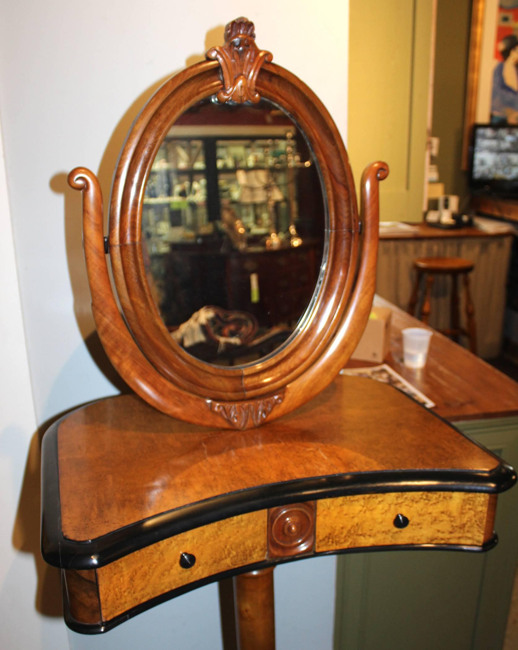 A 19th century Biedermeier two drawer shaving stand, with bird’s-eye maple veneer highlighted by ebonized mahogany, and an oval mirror in a carved mahogany frame. Supported by a columnar flame birch pedestal terminating with three foliate and scroll