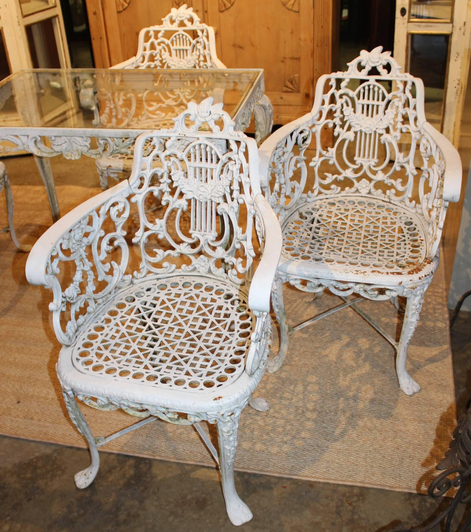 A wonderful heavy cast iron white painted garden set which includes four armchairs and a glass top table, all with scroll decoration and rose crests, along with cabriole legs. Each chair is reinforced with cross stretchers. The set is unsigned, but