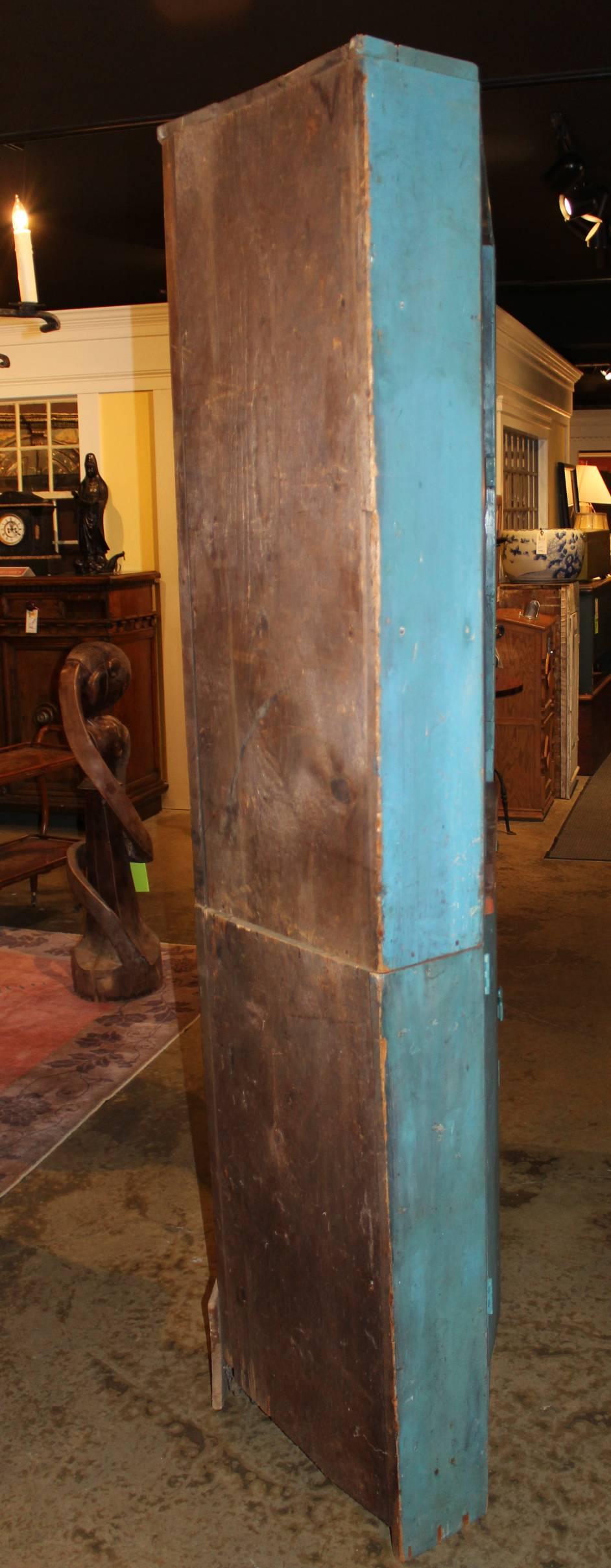 A fine two part wooden corner cupboard in old blue paint, the open upper case with a red wash interior, and two shaped shelves, over a lower case with two doors which open to reveal two interior shelves. Dates to the 19th century, with some repairs