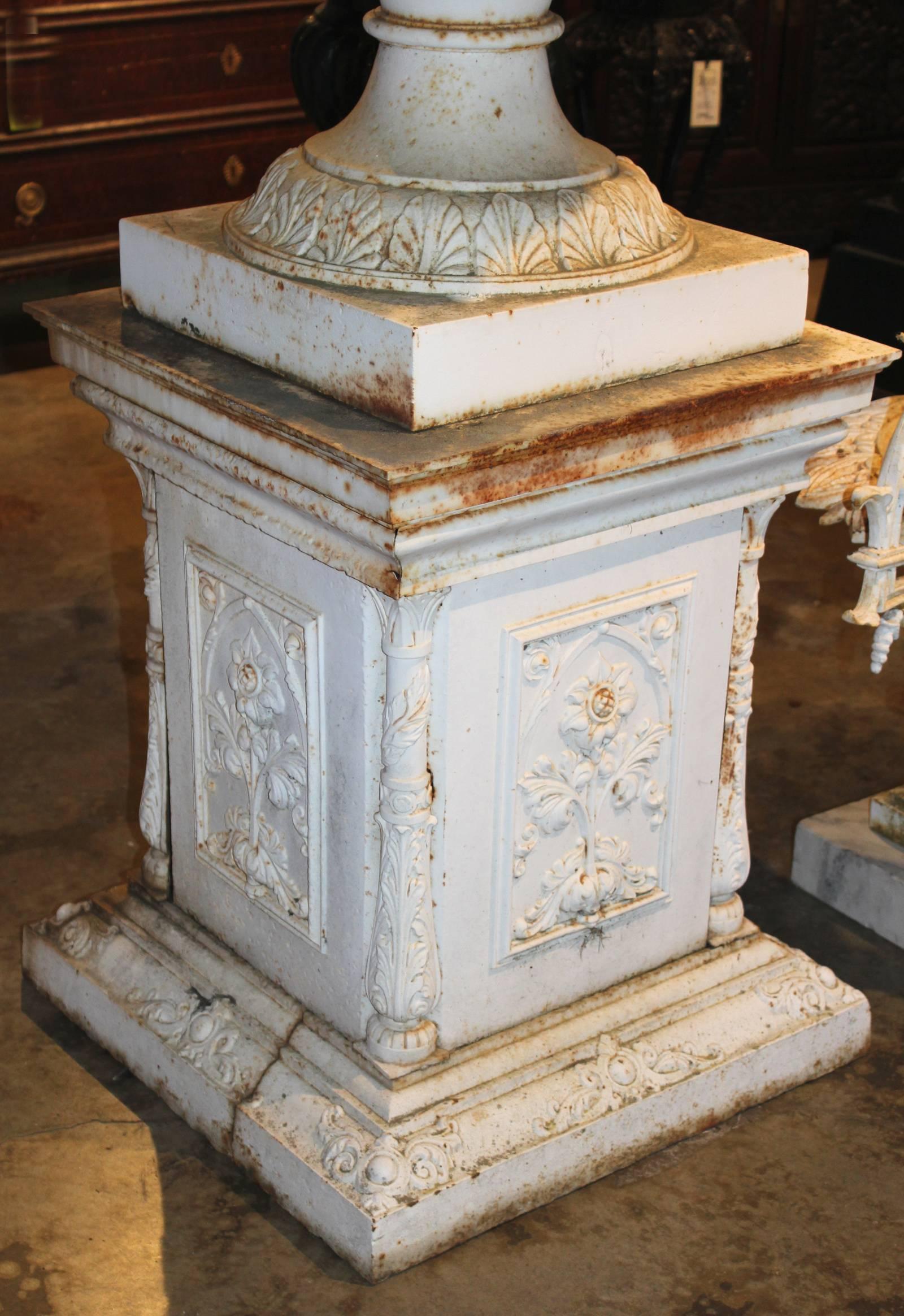 A spectacular pair of white painted Victorian iron urns with lily pad and scallop decorated rims and foliate pattern bases, on iron plinths with foliate Gothic arched panels on all four sides, and foliate quarter columns on each corner. Very good
