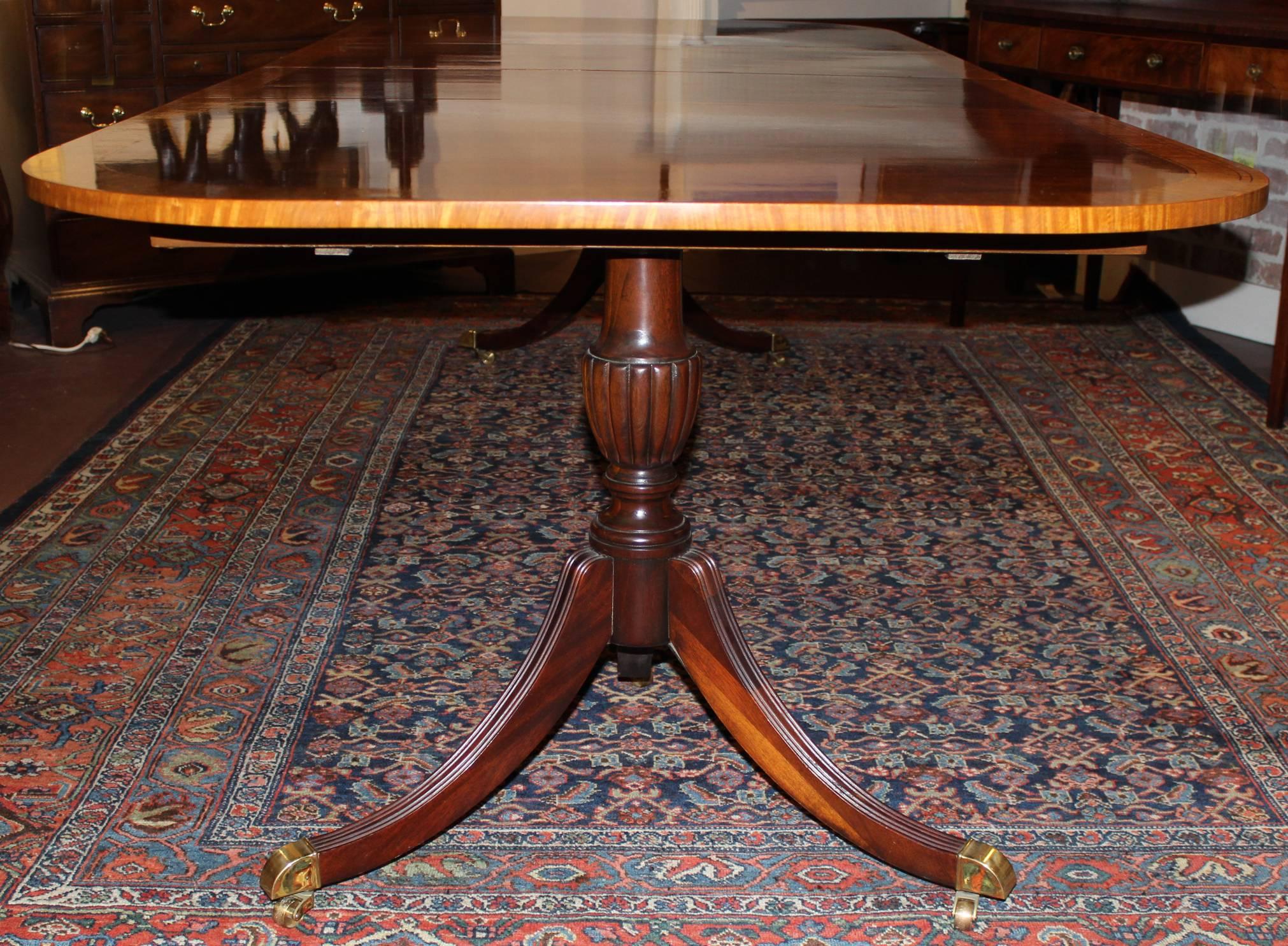 A splendid rectangle mahogany veneered double pedestal dining table, its top with rounded corners and a satinwood double-banded border, each pedestal with three down swept legs terminating with brass capped casters. Two leaves included. Dates to the